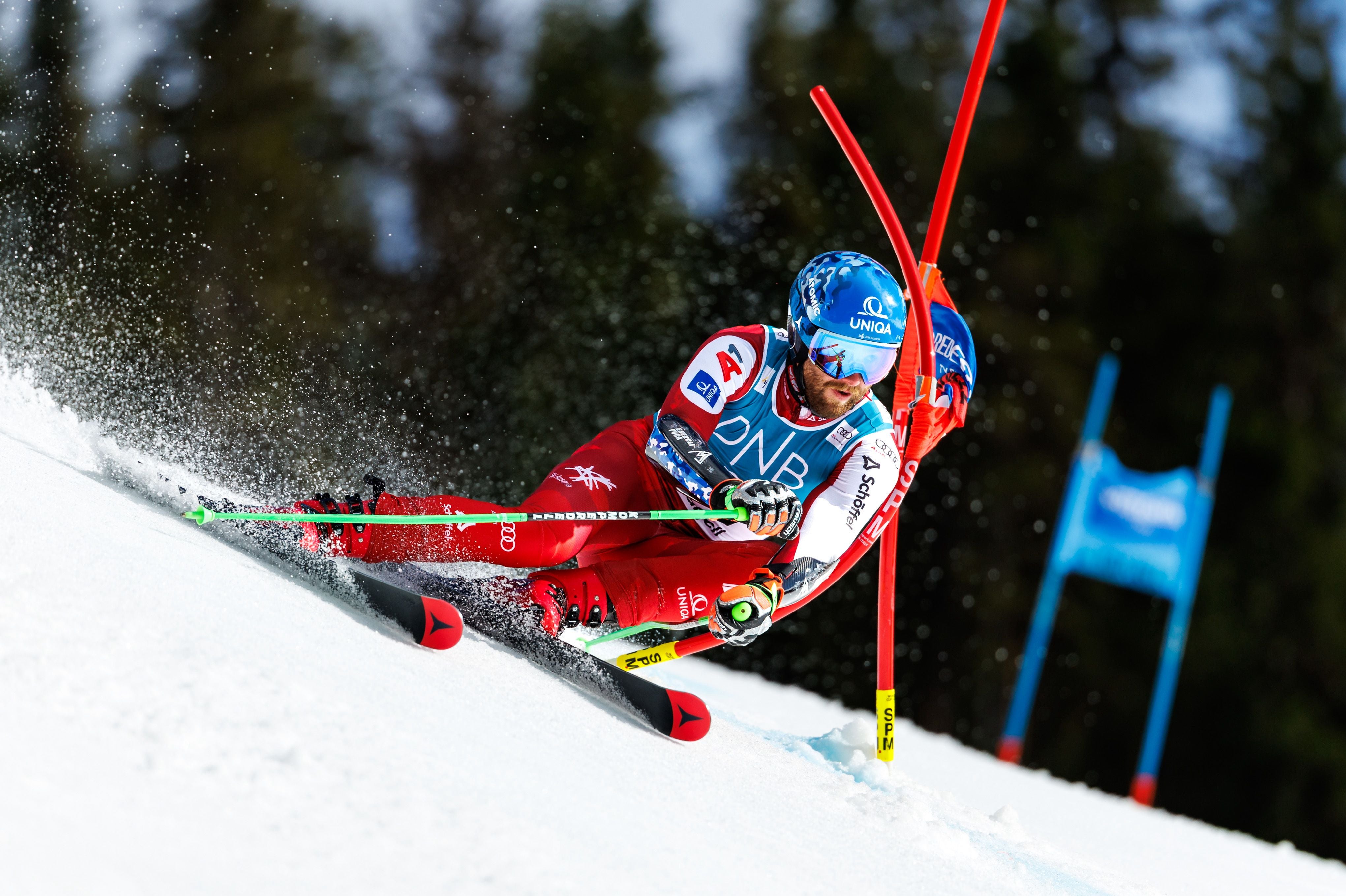 ÖSV-Ass Marco Schwarz zieht im roten Rennanzug dynamisch eine scharfe Kurve um ein rotes Slalomtor auf einer präparierten Piste.
