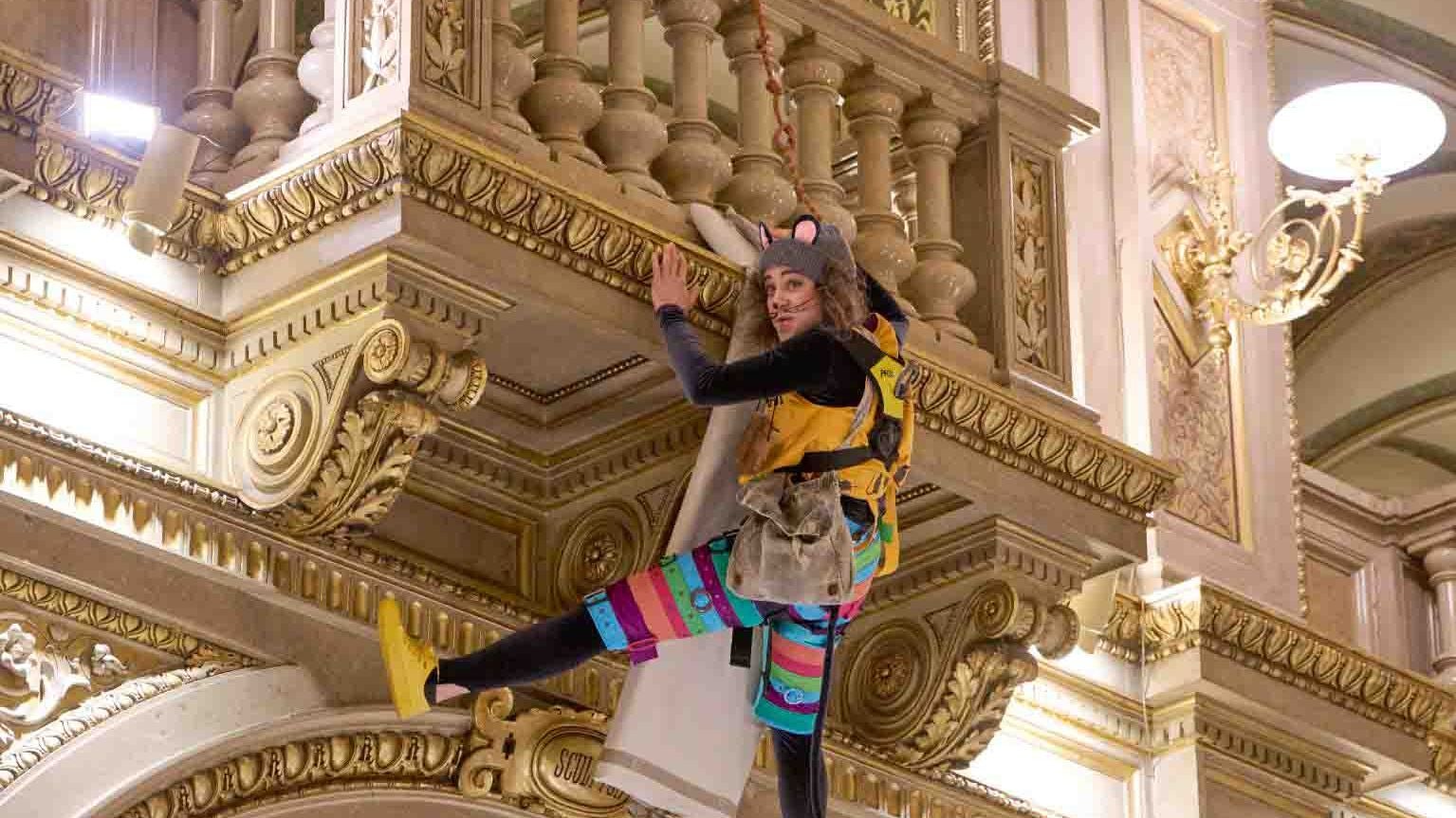 Eine Person im bunten Kostüm hängt verkleidet als Maus in einem prunkvollen Opernhaus.