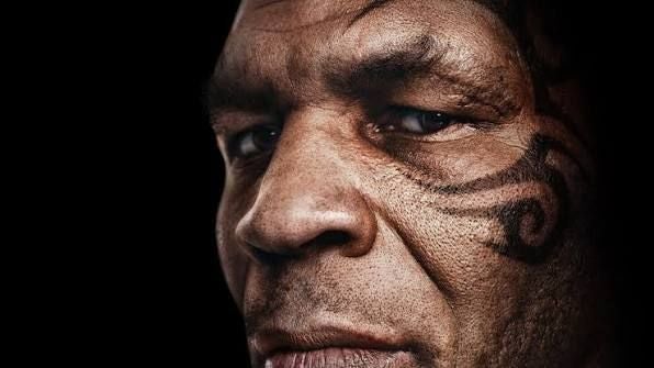 Nahaufnahme eines Mannes mit Gesichtstattoo und ernstem Blick, dunkler Hintergrund.