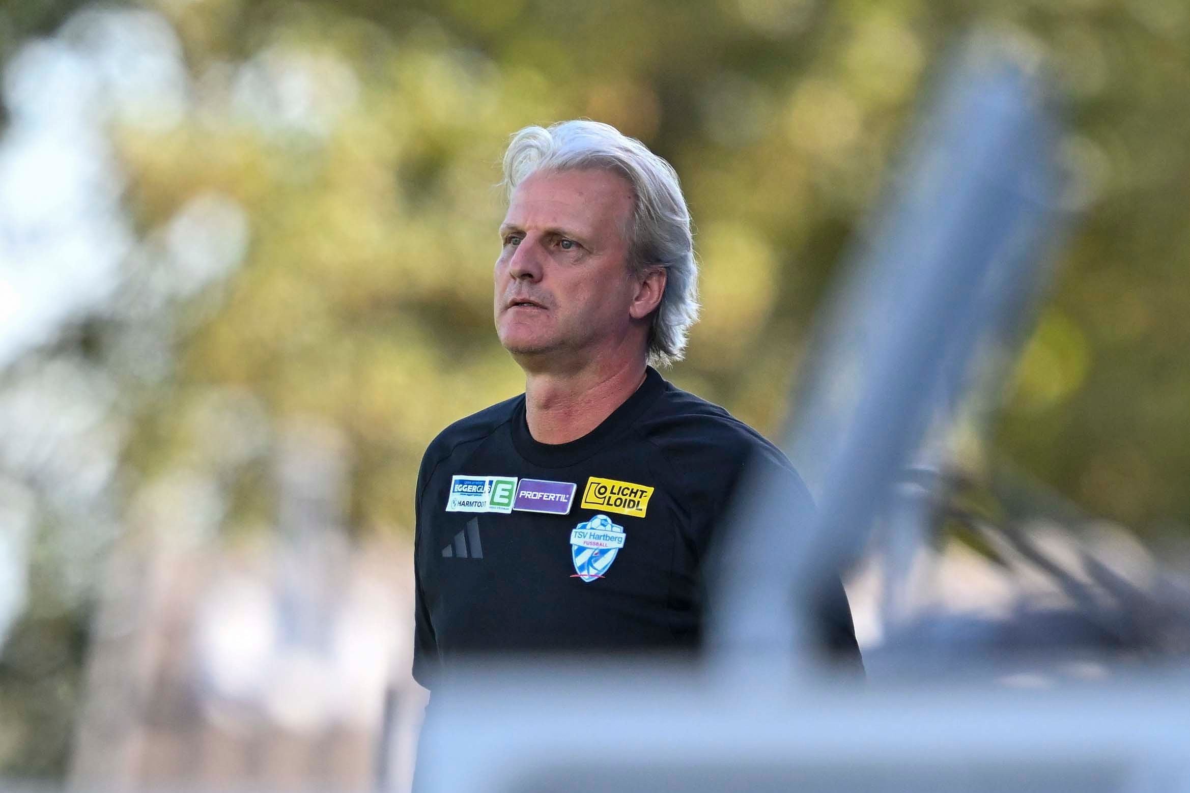 Hartberg-Trainer Markus Schopp steht im schwarzen Trainingsshirt mit Vereinswappen auf dem Sportplatz und blickt aufmerksam zur Seite.