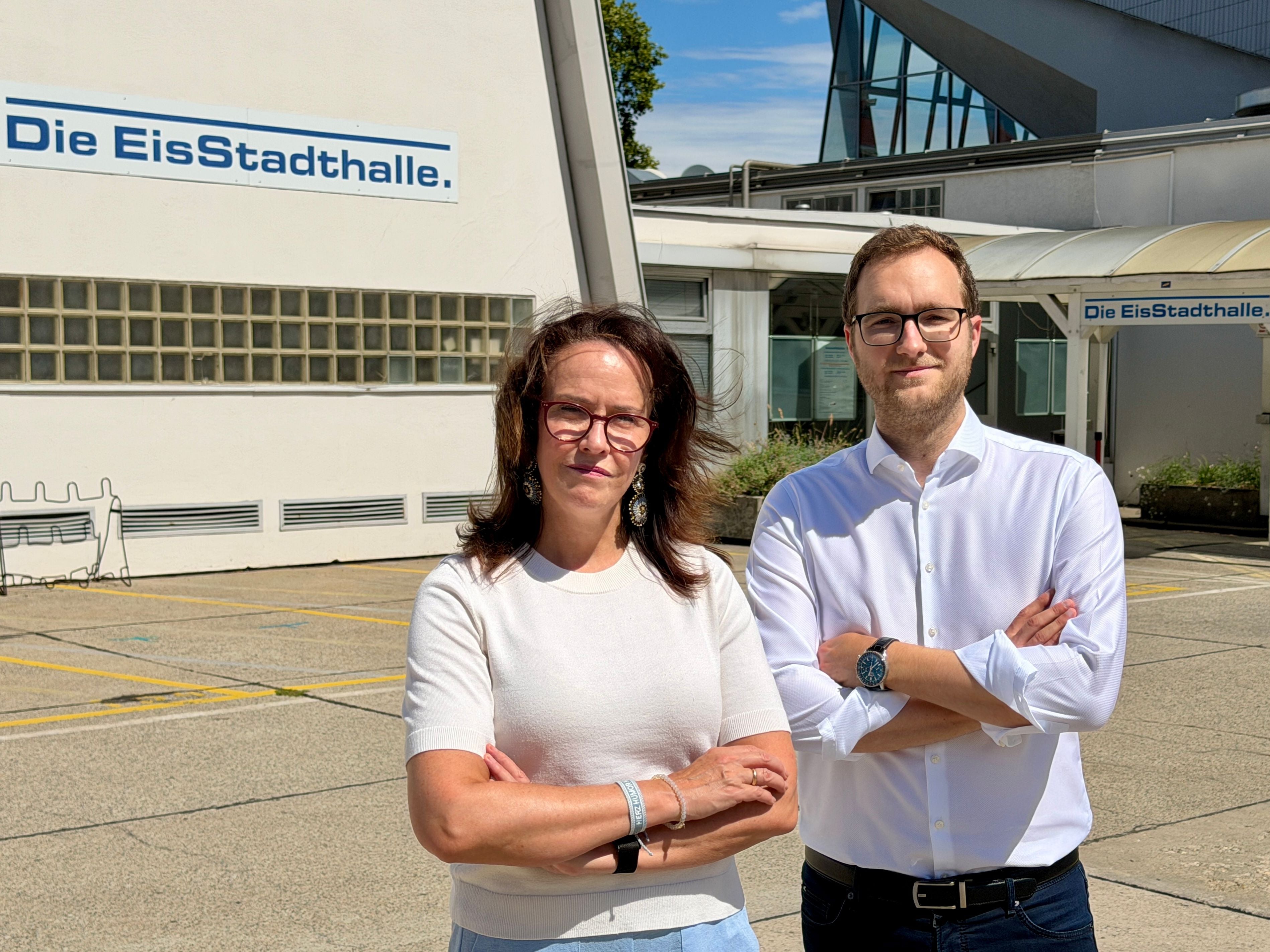 Eine Frau und ein Mann stehen mit verschränkten Armen vor der EisStadthalle im Freien.