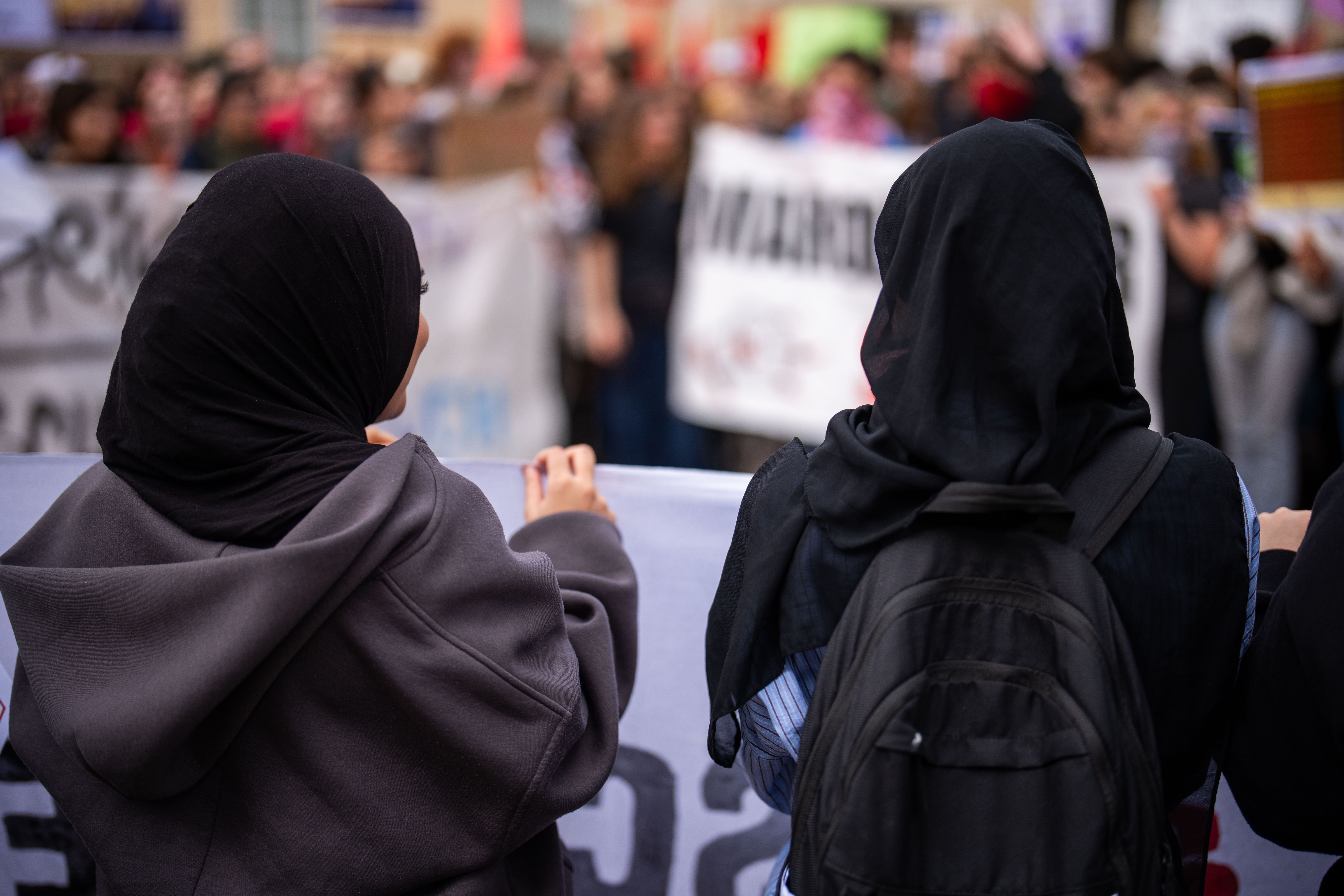 Zwei Frauen mit Kopftuch bei einer Kundgebung gegen das Kopftuchverbot, Menschen und Banner im Hintergrund.