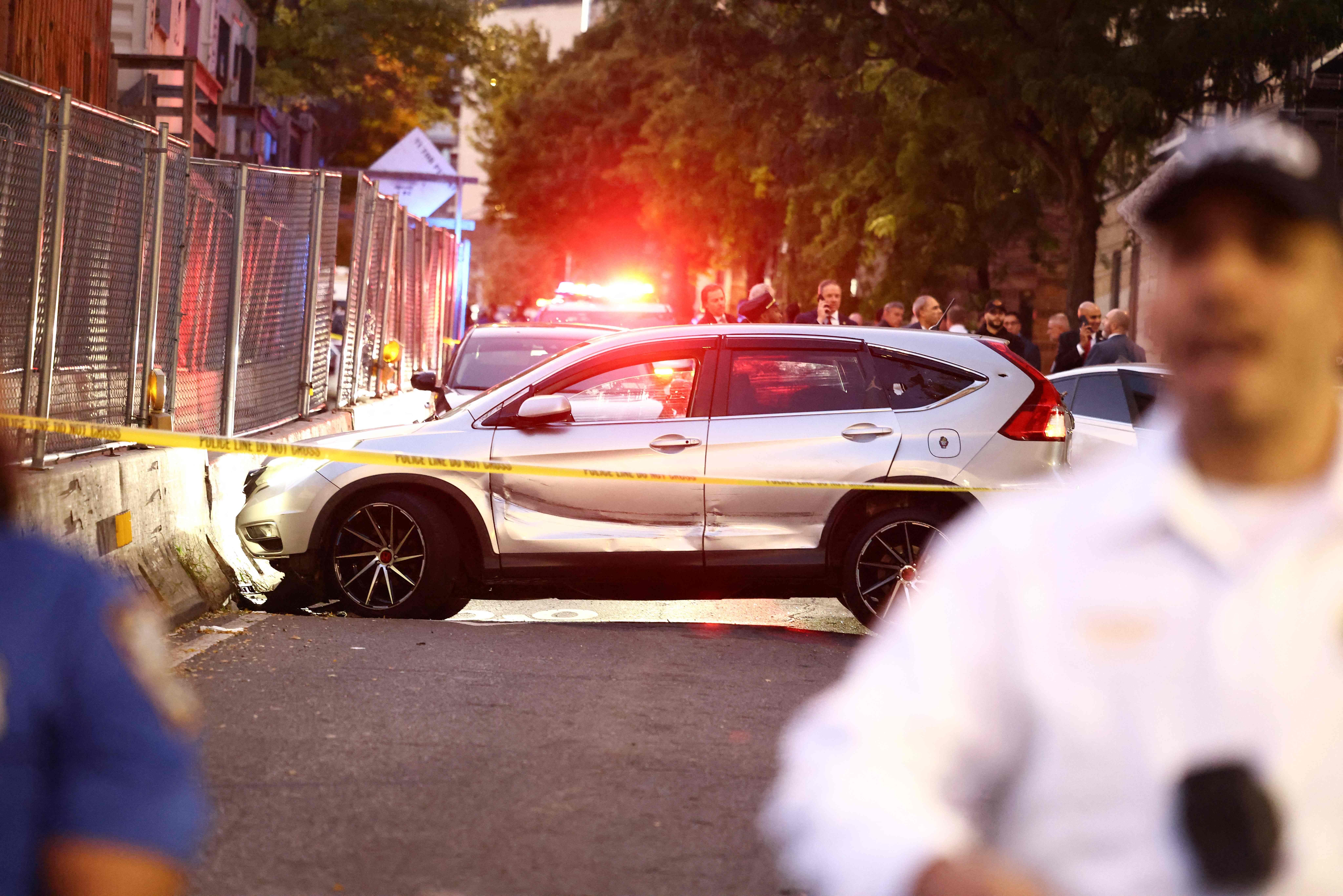 Polizeiauto mit Absperrband und Blaulicht, Menschen stehen im Hintergrund auf einer Straße.