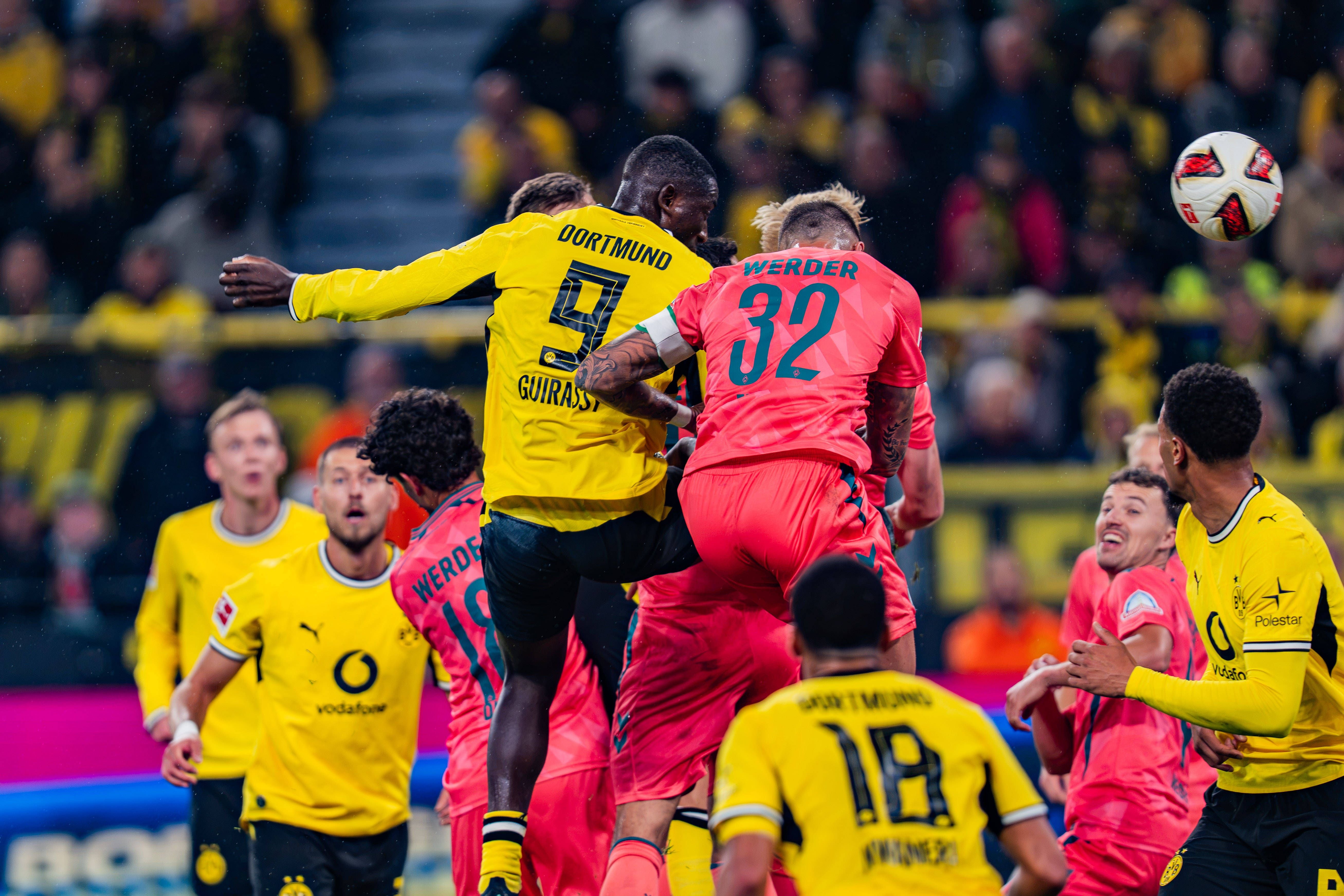 Fußballspiel zwischen Dortmund und Bremen, Spieler springen zum Kopfball, Zuschauer im Hintergrund.