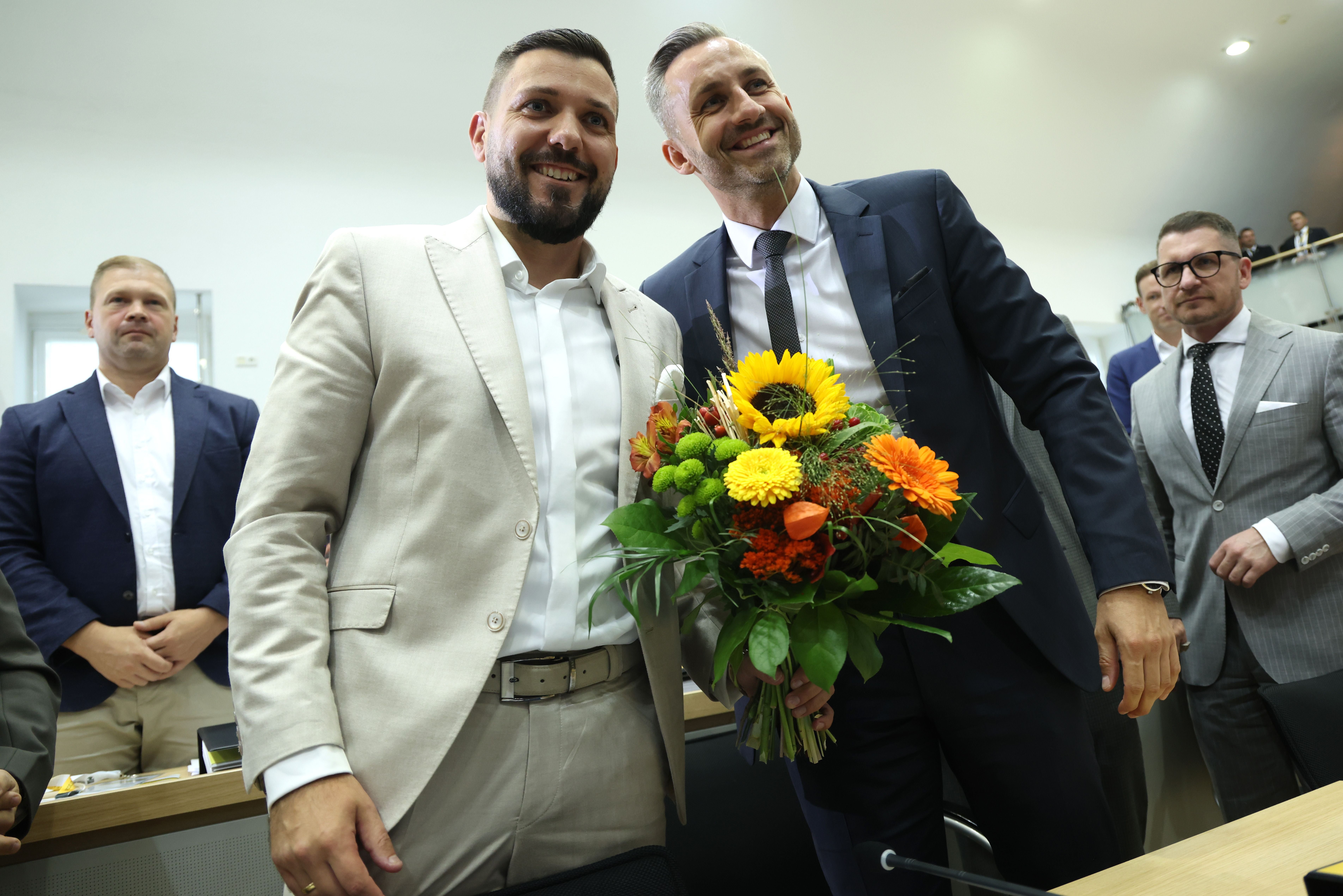 Zwei Männer im Anzug posieren lächelnd, einer hält einen Blumenstrauß, mehrere Männer im Hintergrund.
