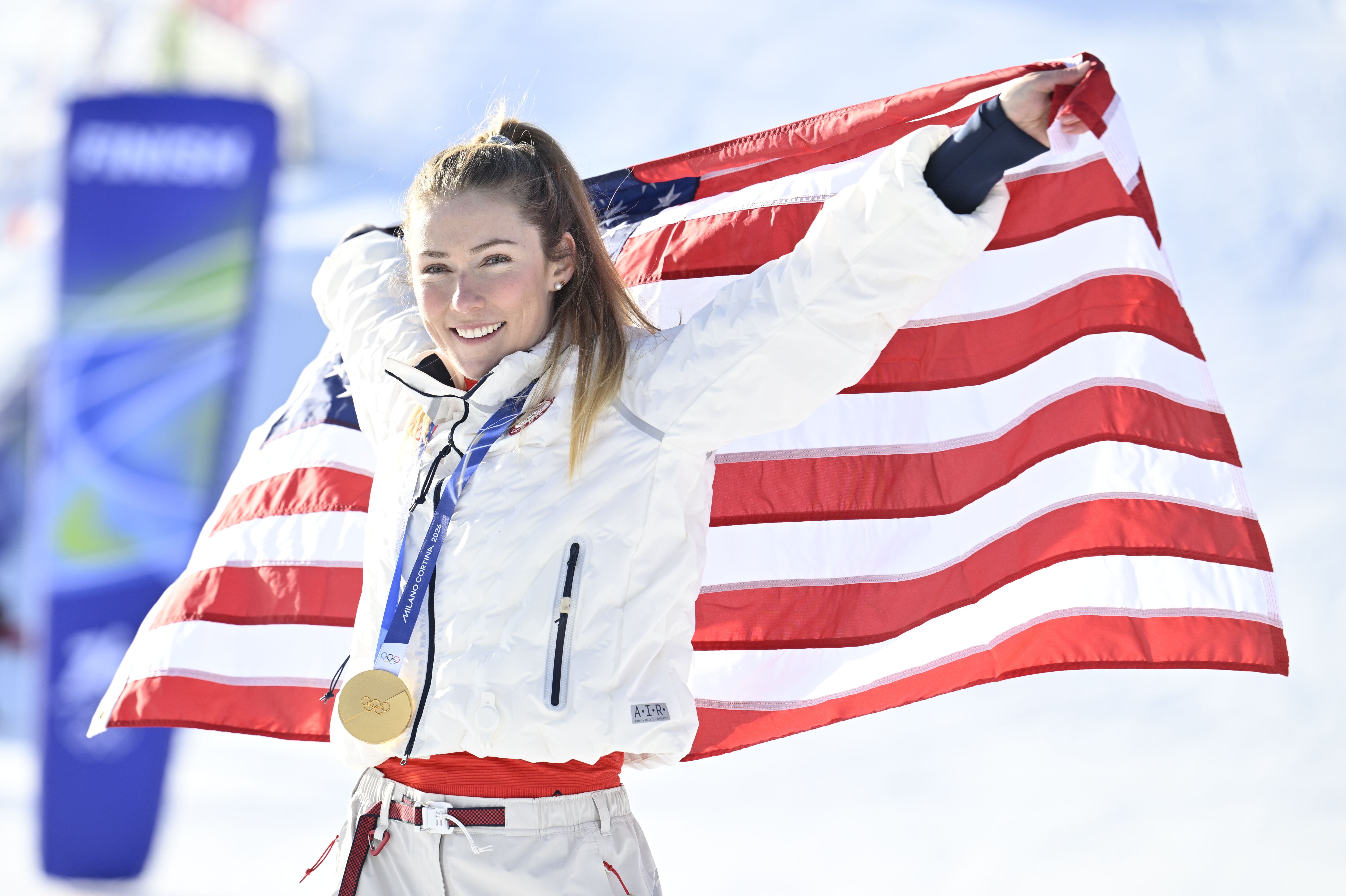 Eine Skifahrerin mit Goldmedaille hält die US-Flagge und lächelt im Schnee.