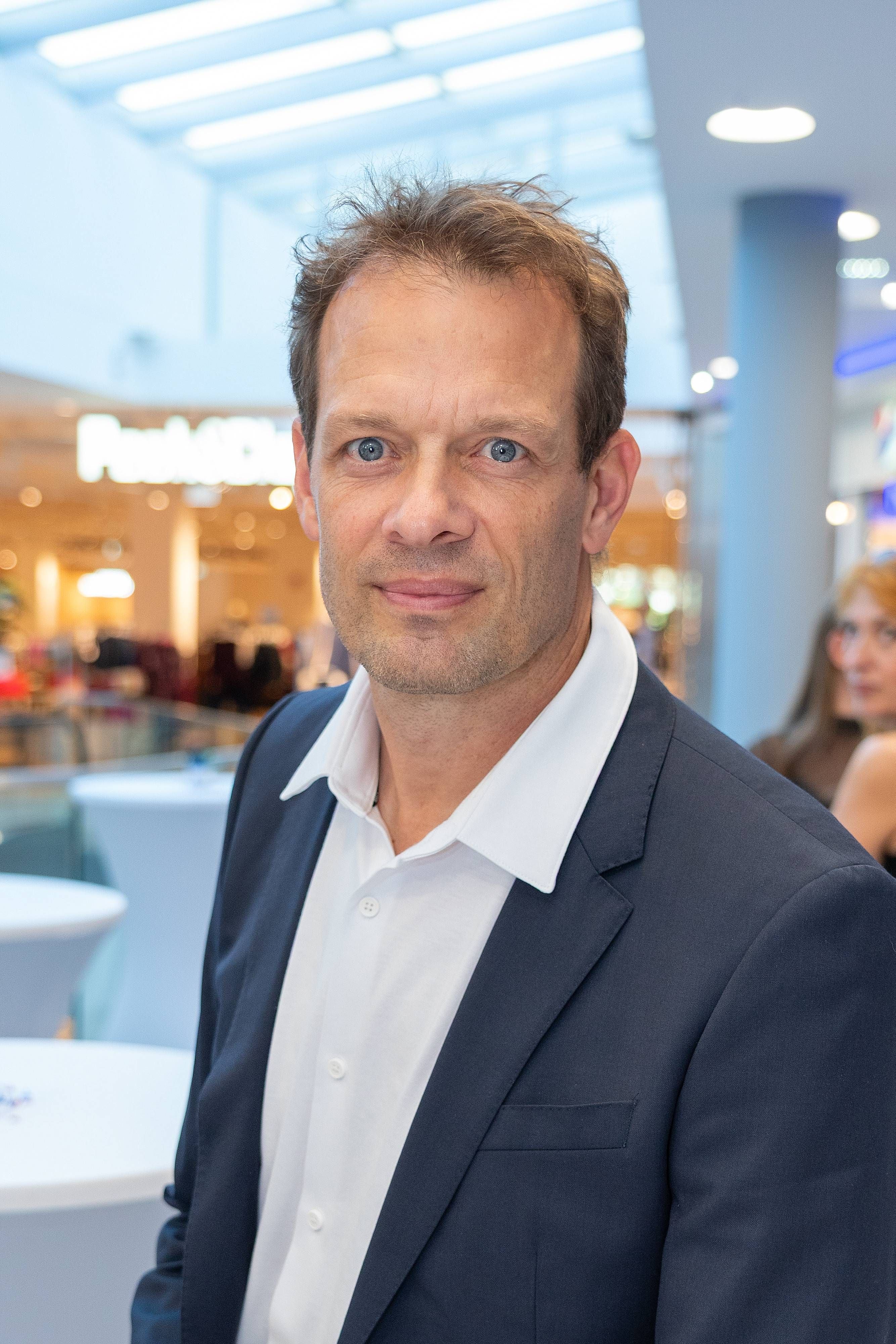 Ein Mann im Anzug steht in einem modernen, hellen Einkaufszentrum und blickt in die Kamera.