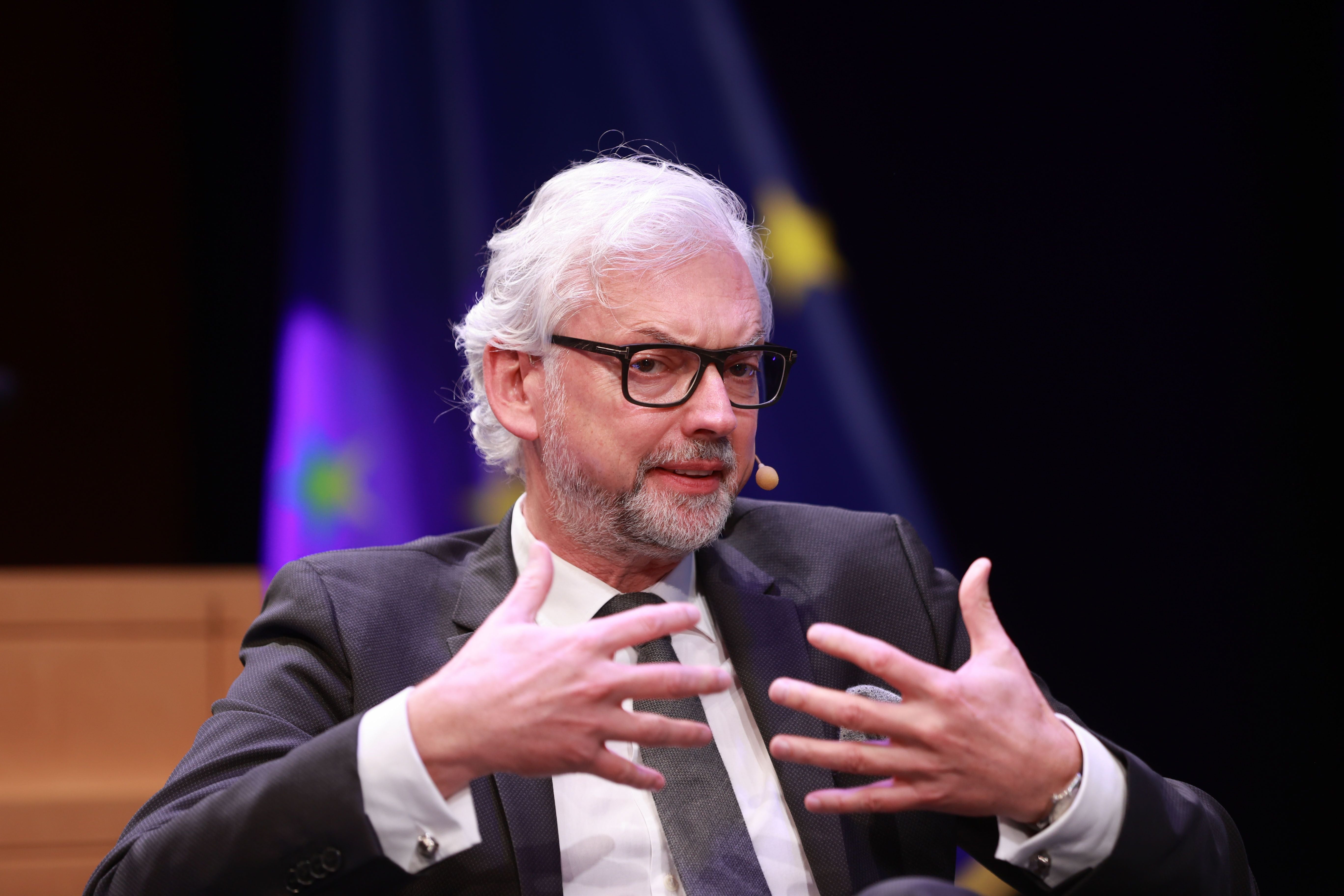 Ein Mann mit Brille spricht gestikulierend bei einer Konferenz, im Hintergrund EU-Sterne.