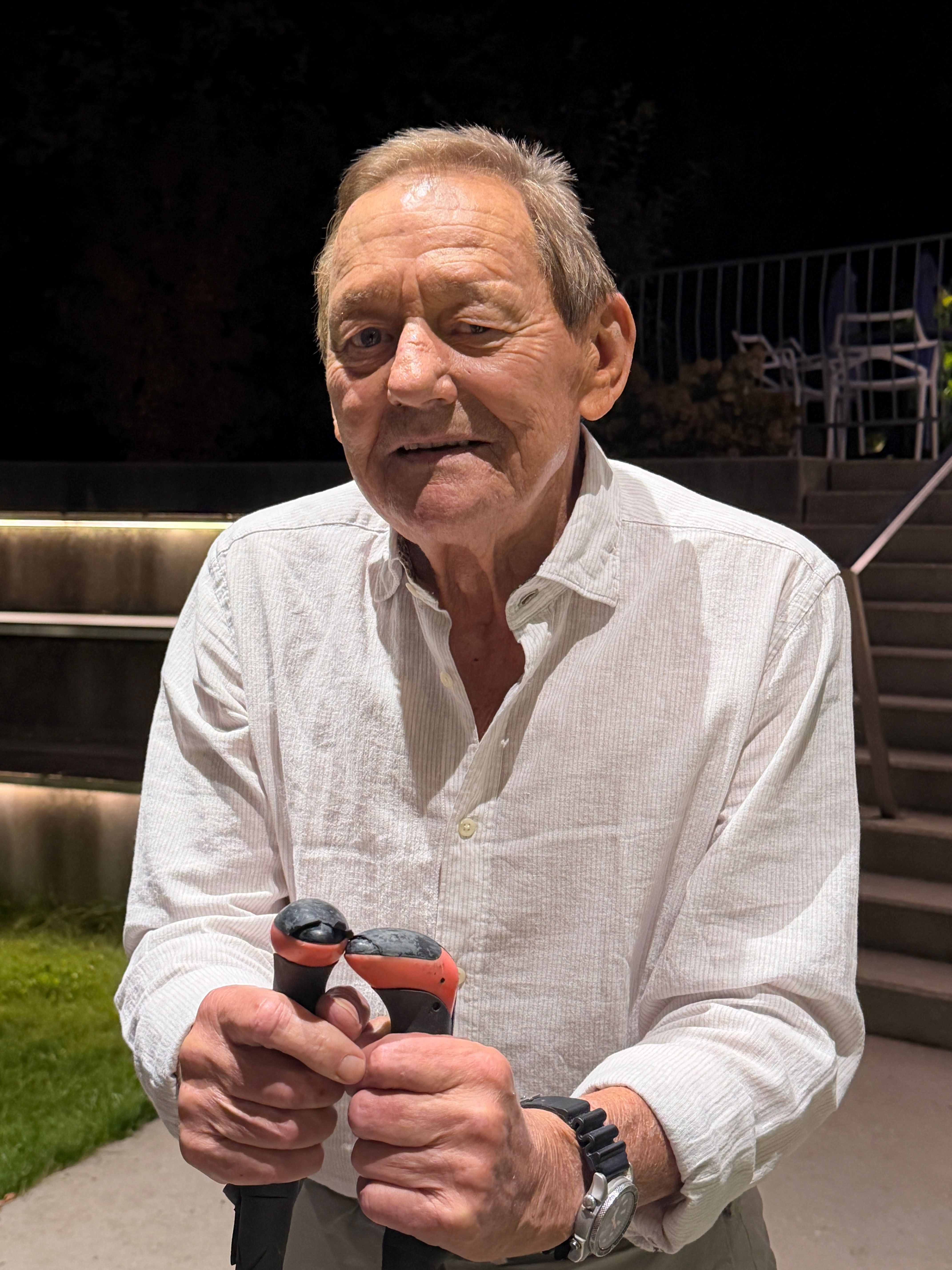 Älterer Mann in weißem Hemd hält zwei Gehstöcke, steht draußen bei Nacht vor einer Treppe.