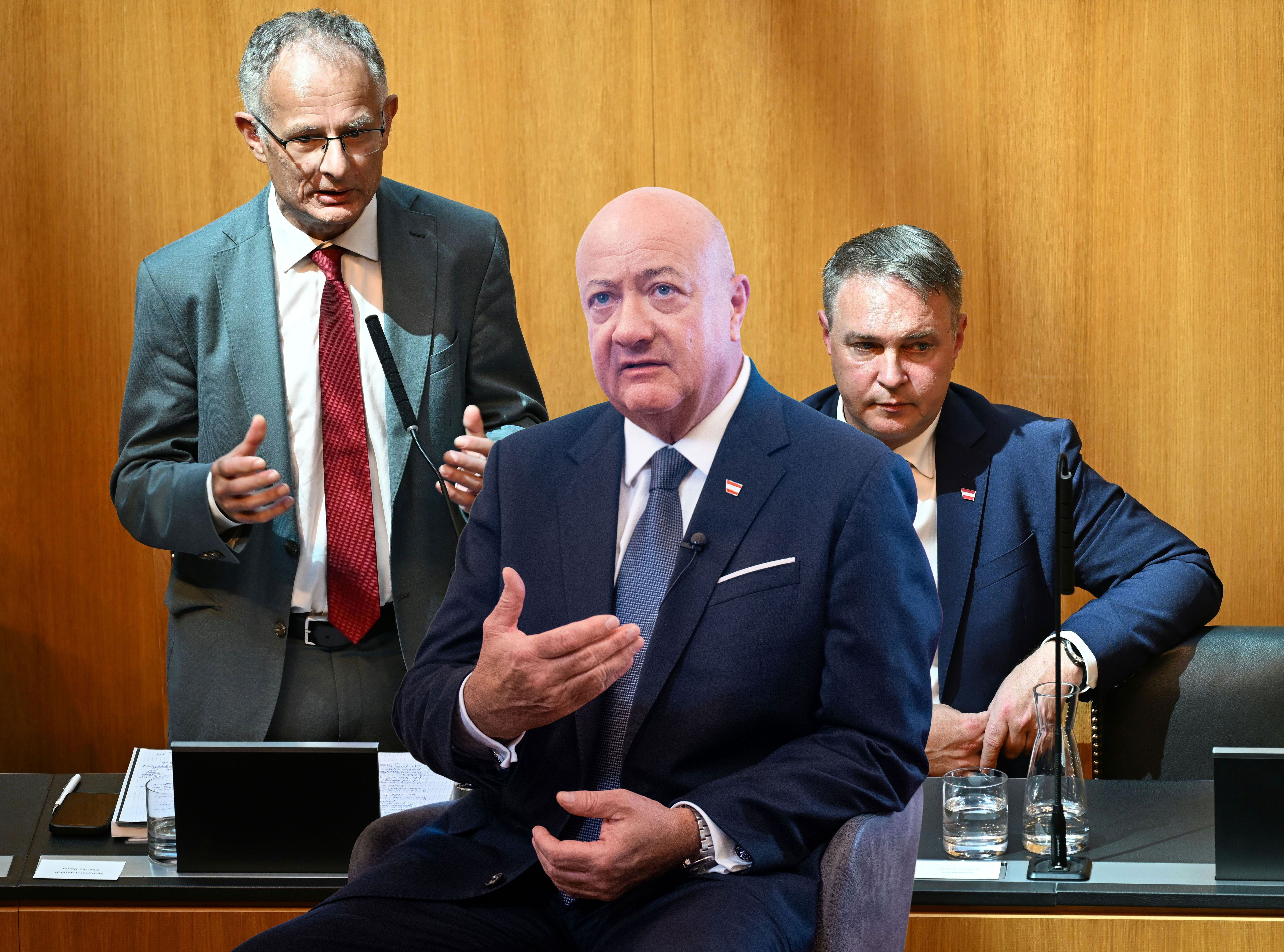 Drei Männer in Anzügen diskutieren in einem Raum mit hölzerner Wand und Mikrofonen.