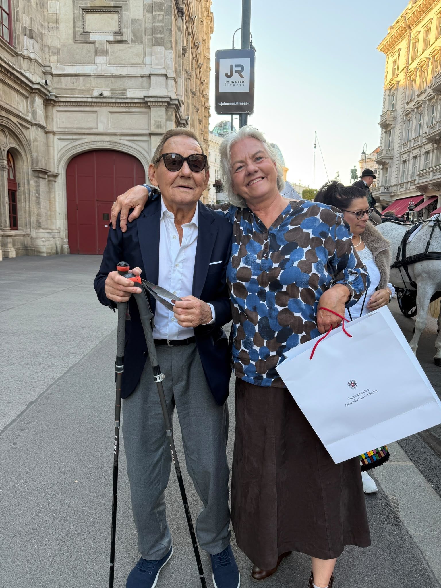 Ein älterer Mann mit Sonnenbrille und eine Frau posieren lächelnd auf einer Stadtstraße.