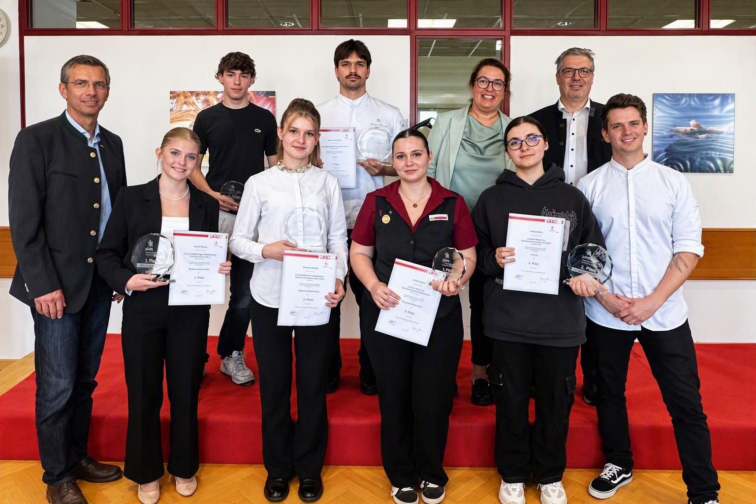 Eine Gruppe von zehn Personen posiert auf einer Bühne, einige halten Urkunden und Glastrophäen.