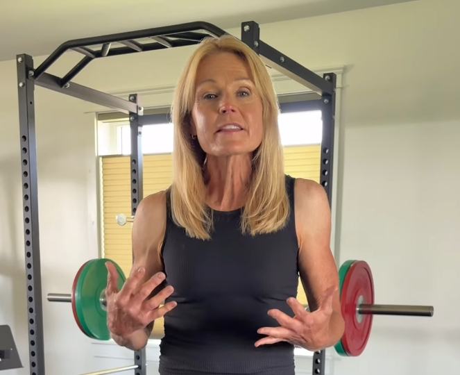 Frau spricht in einem Fitnessraum vor einer Langhantel mit Gewichtsscheiben.