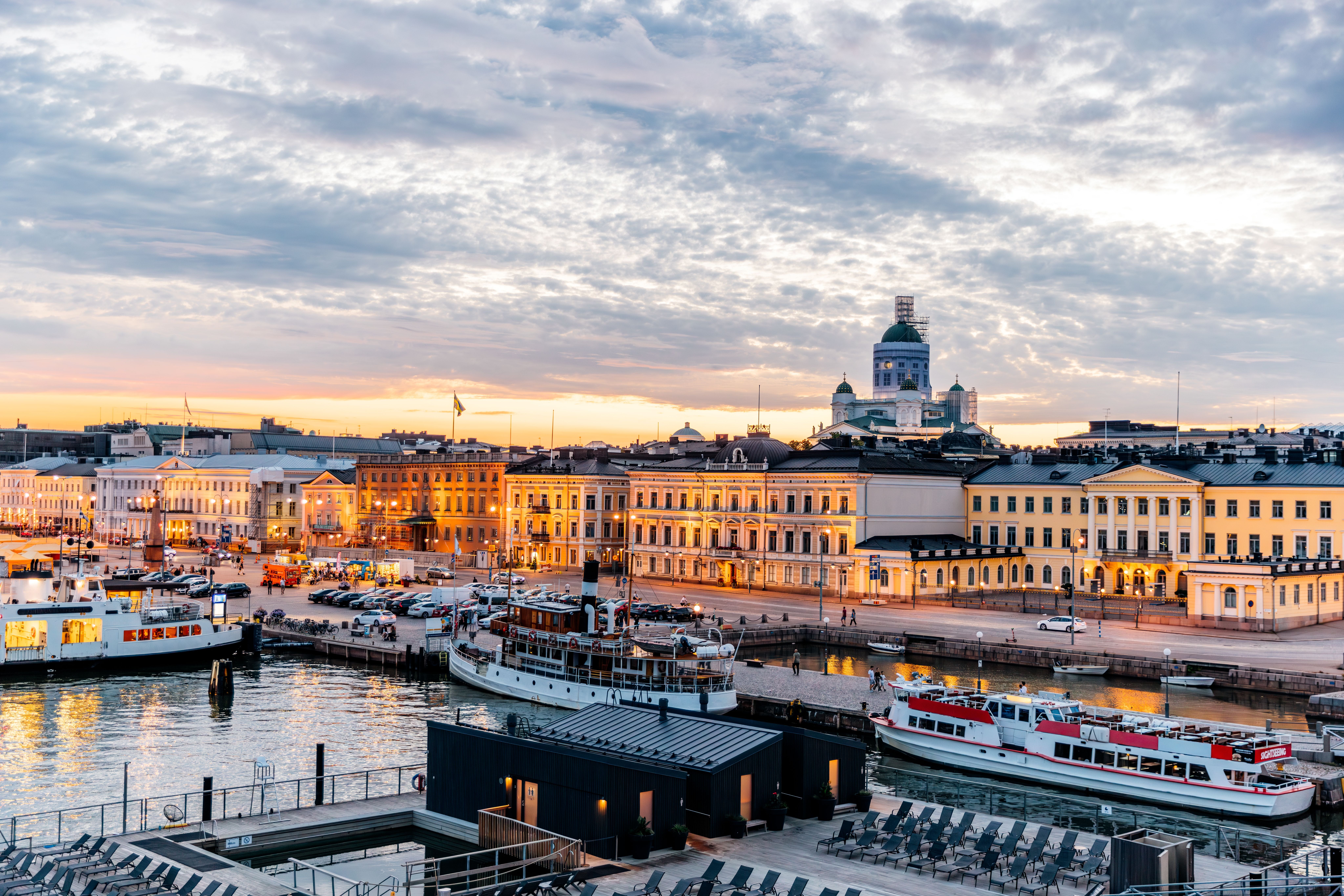 Hafen von Helsinki bei Sonnenuntergang mit Gebäuden, Booten und dem Dom im Hintergrund.