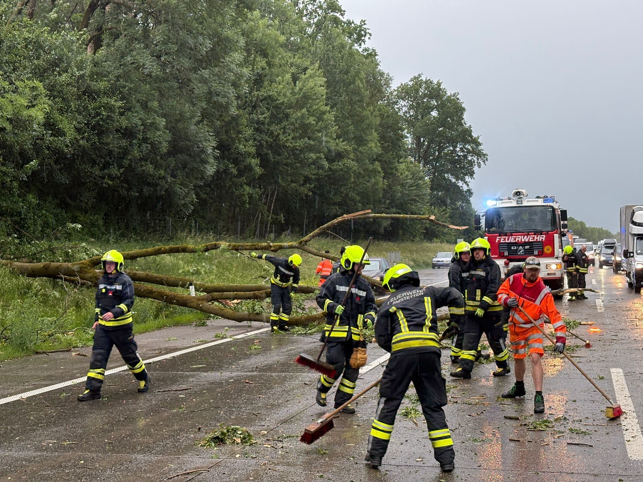 Feuerwehr entfernt nach Sturm umgestürzte Bäume von nasser Autobahn, Einsatzfahrzeuge im Hintergrund.