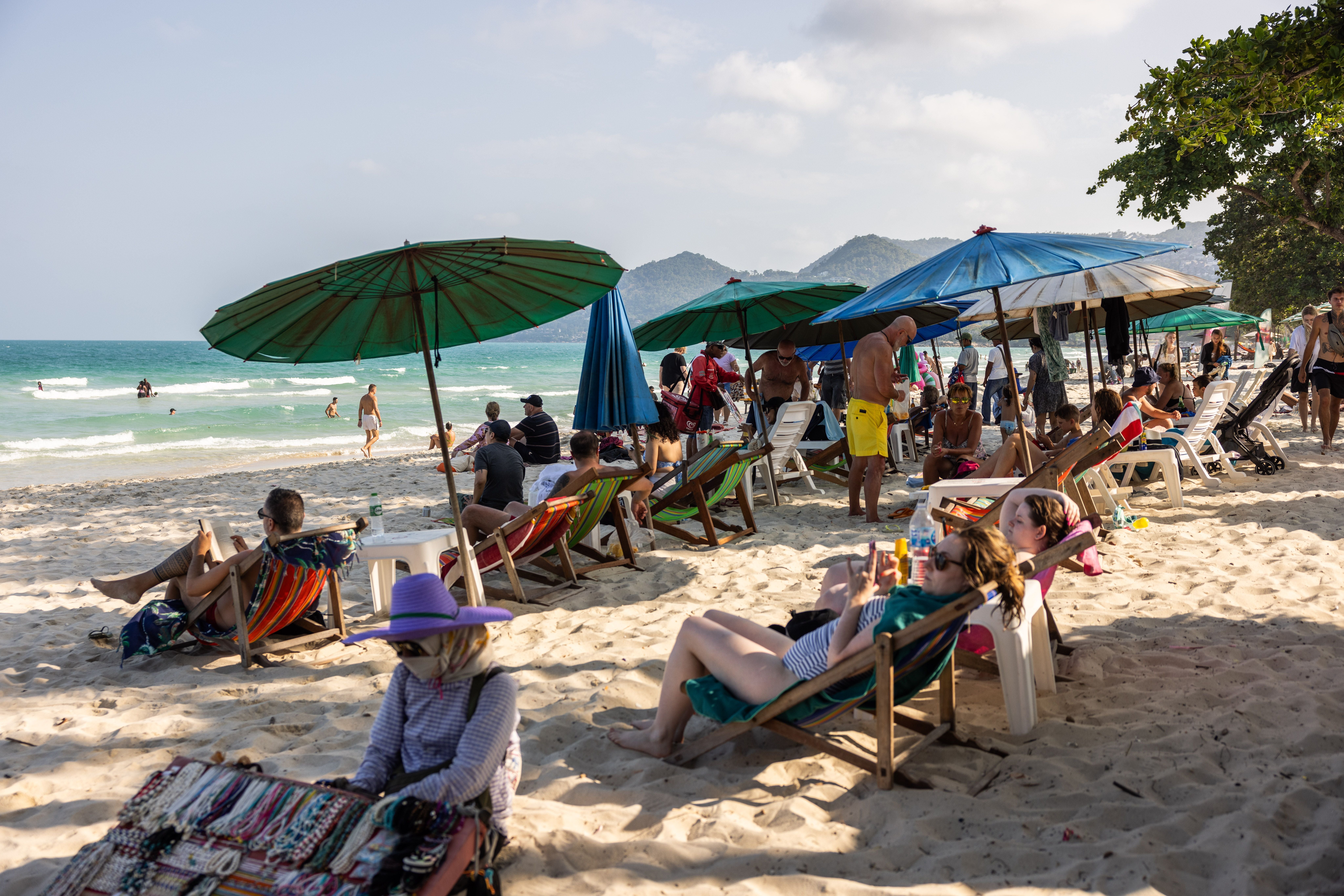Touristen entspannen unter Sonnenschirmen an einem belebten Sandstrand mit Meer im Hintergrund.
