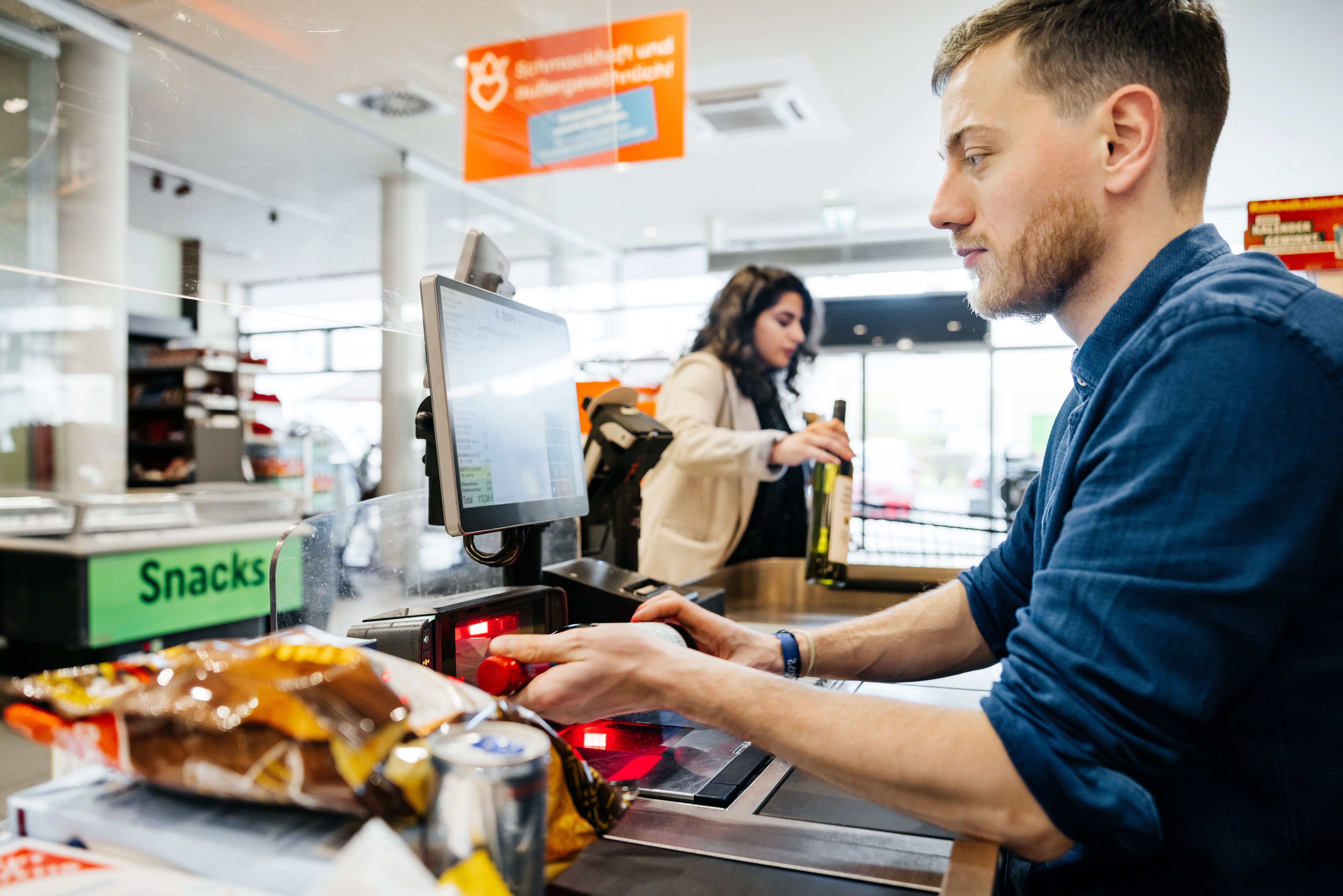 Supermarktmitarbeiter scannt Einkäufe an der Kasse, Kundin legt Produkte aufs Band.