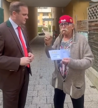 Zwei Männer unterhalten sich draußen, einer hält einen Briefumschlag in der Hand.
