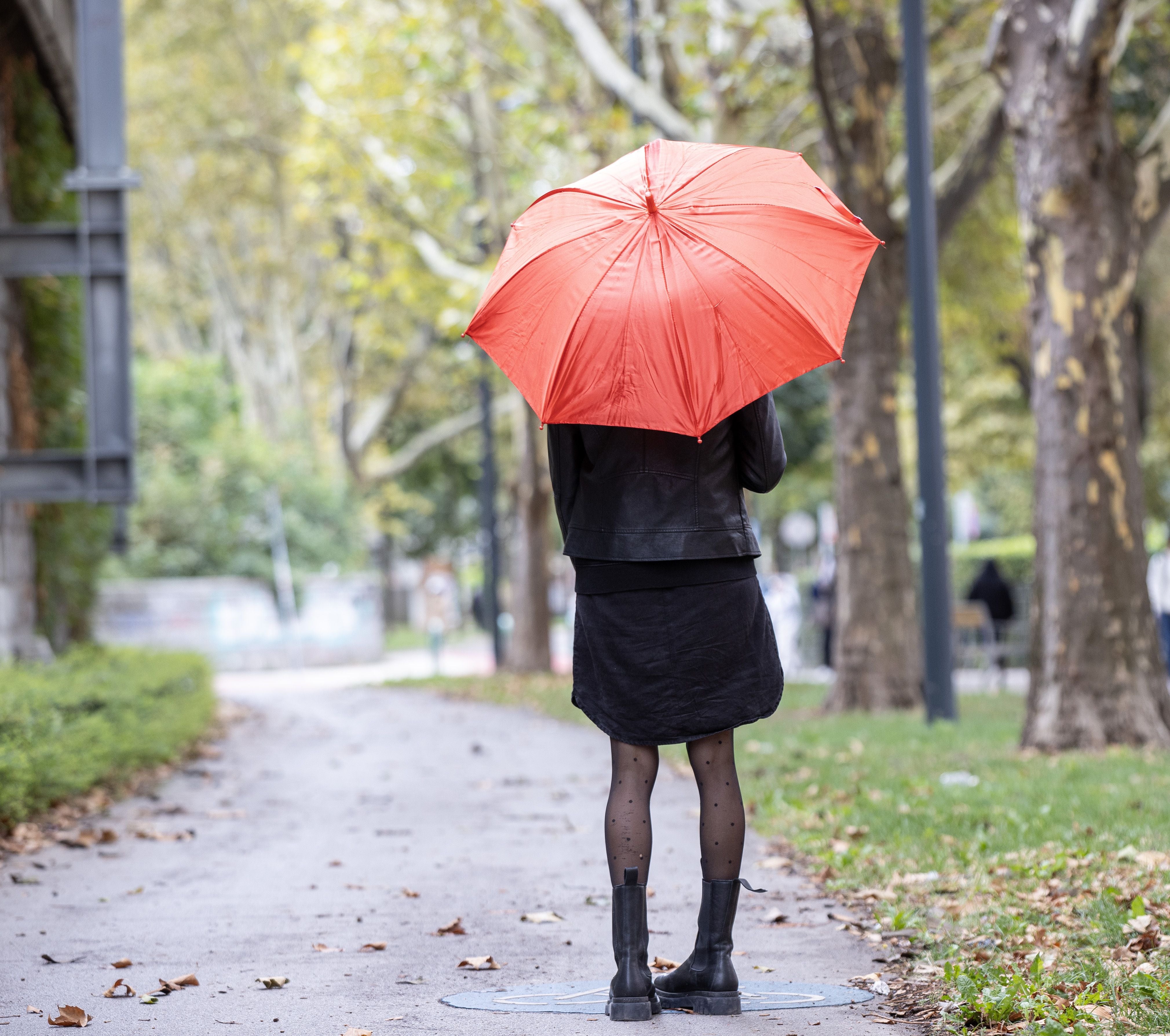Frau mit rotem Regenschirm steht auf einem Weg in einem Park, Bäume im Hintergrund.