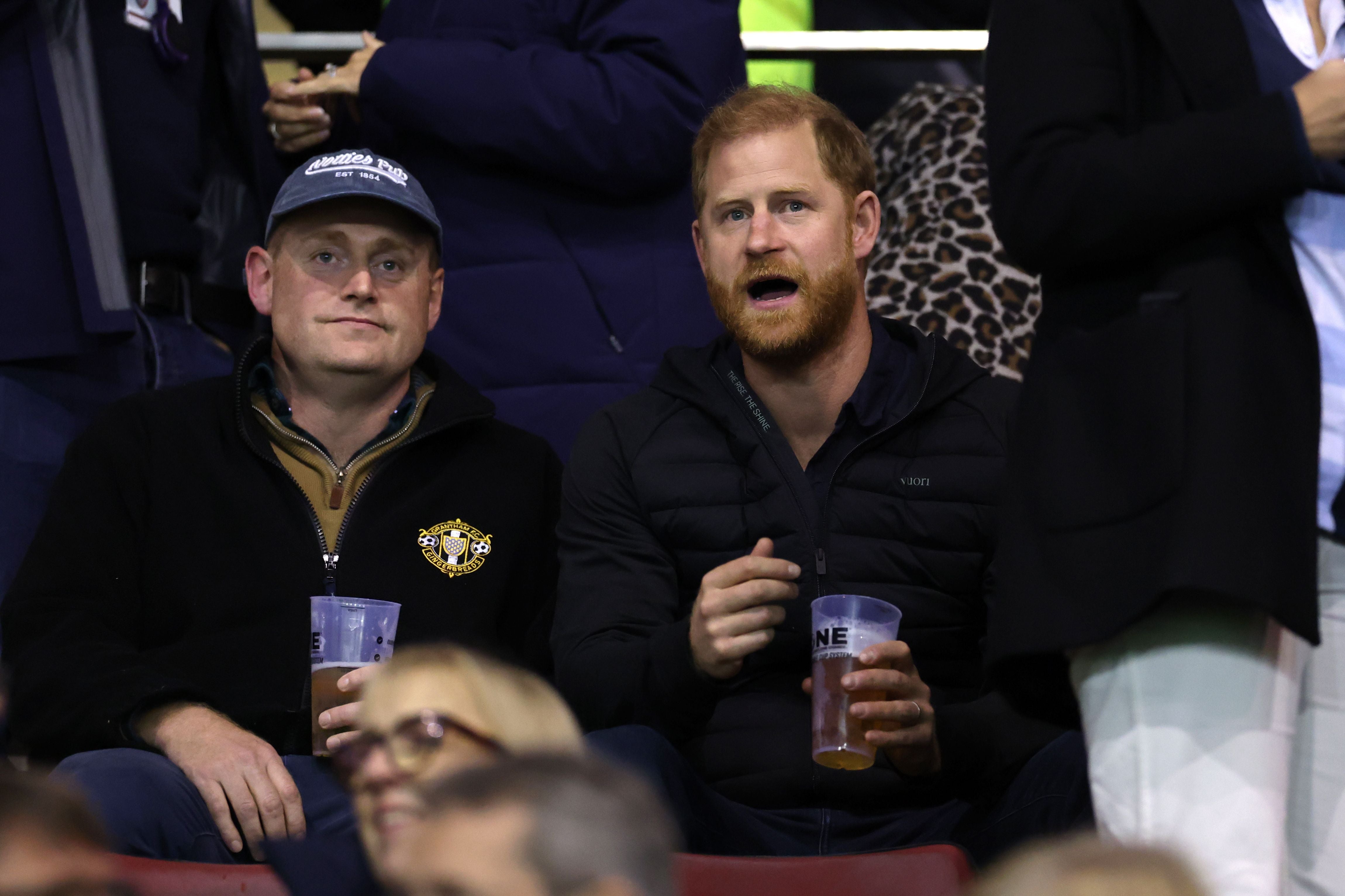Zwei Männer sitzen auf der Tribüne eines Rugby-Stadions und halten Plastikbecher mit Getränken.