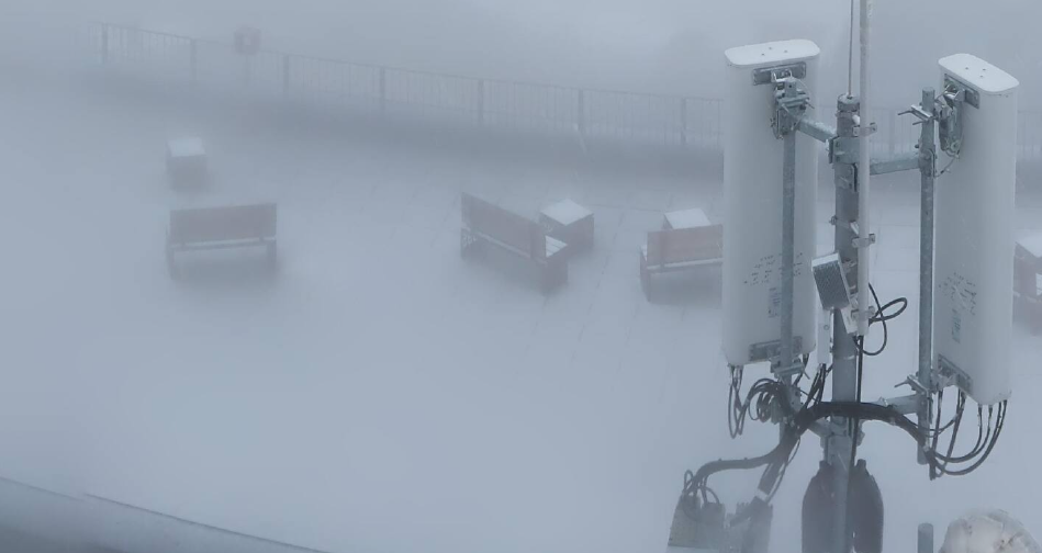 Neblige Szene mit Funkmast, Bänken und Tisch auf einer Terrasse, kaum Sicht durch dichten Nebel.
