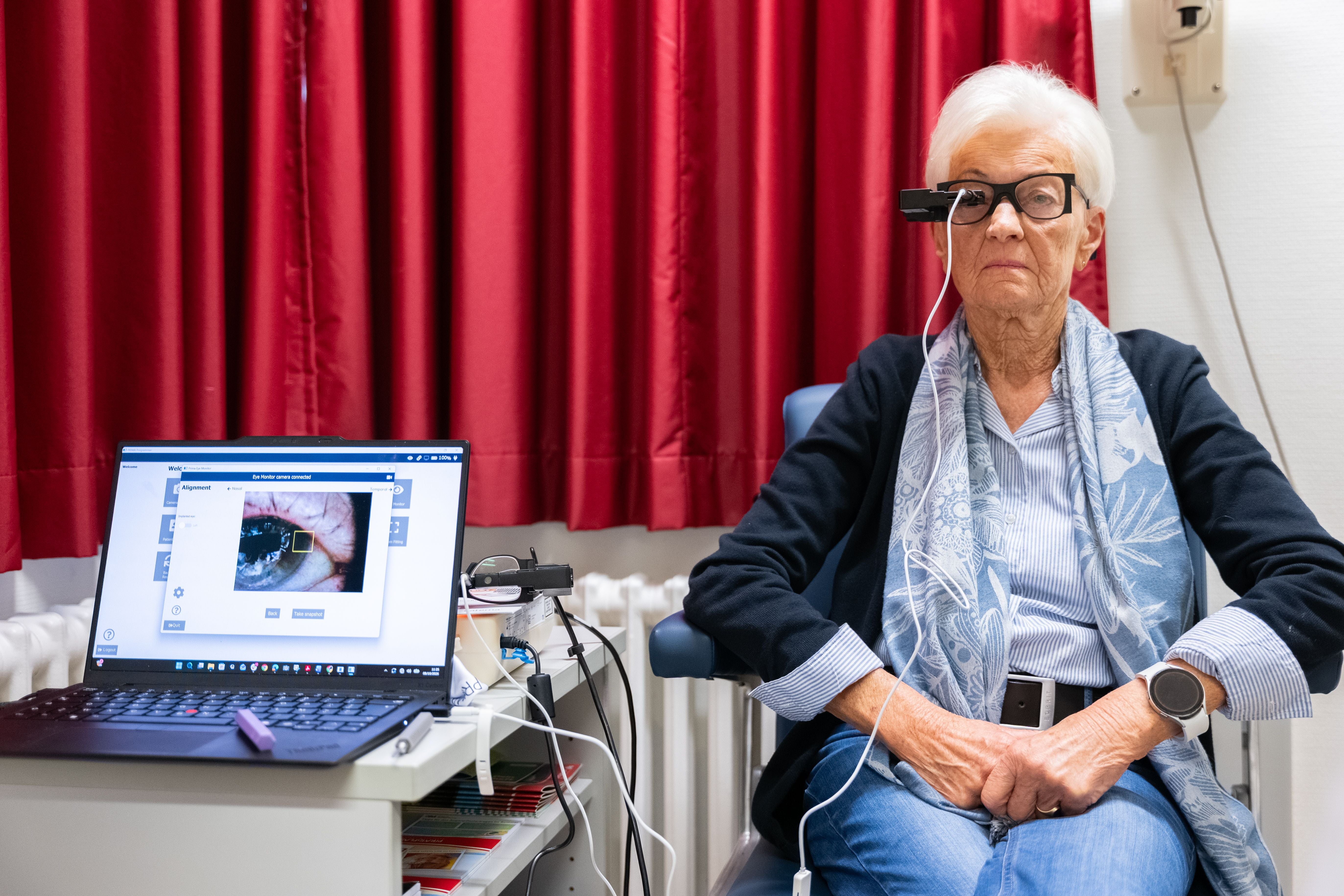 Ältere Frau mit Spezialbrille sitzt vor Laptop, vermutlich zur Netzhautuntersuchung.