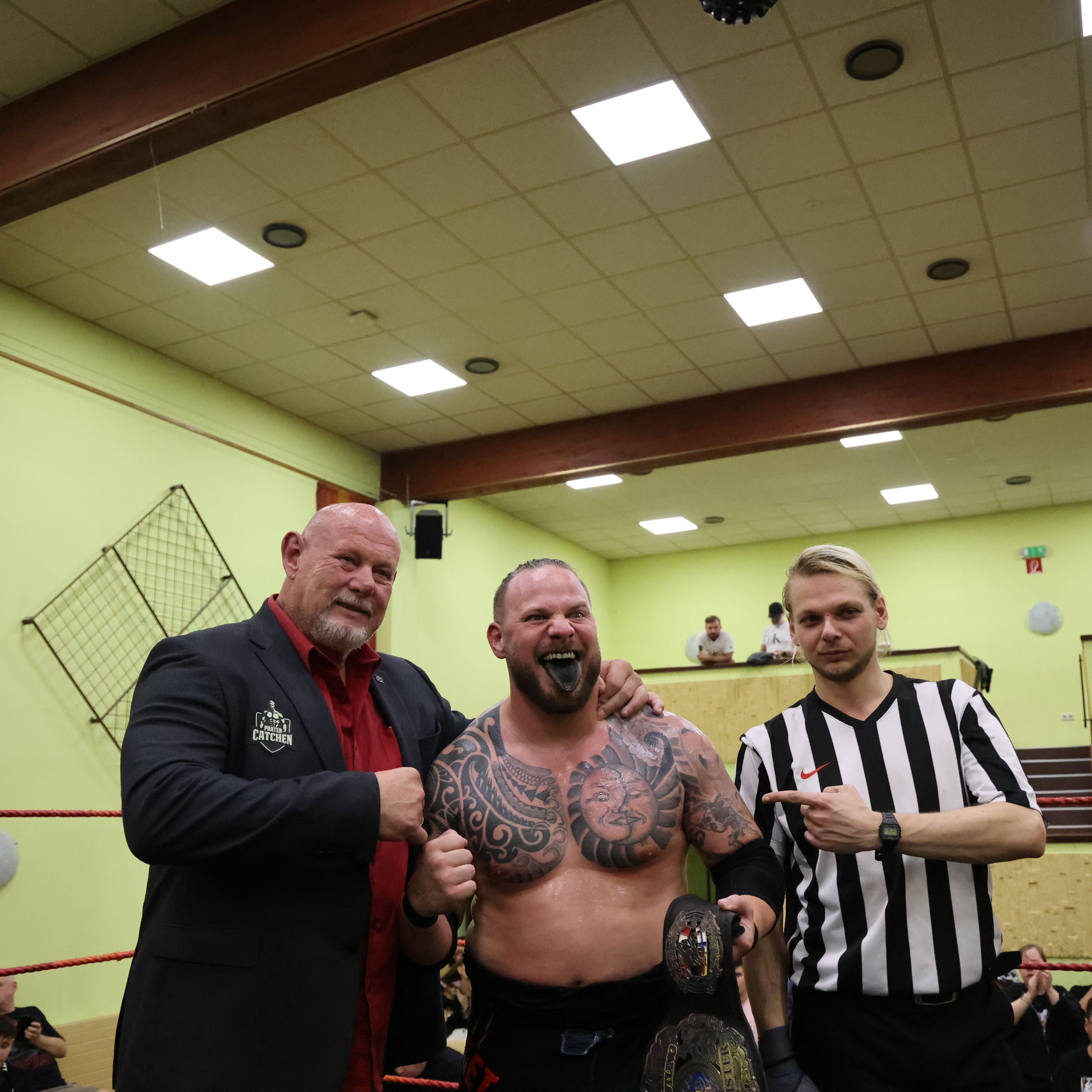 Drei Männer stehen im Wrestling-Ring, einer hält einen Champion-Gürtel, der andere ist Schiedsrichter.