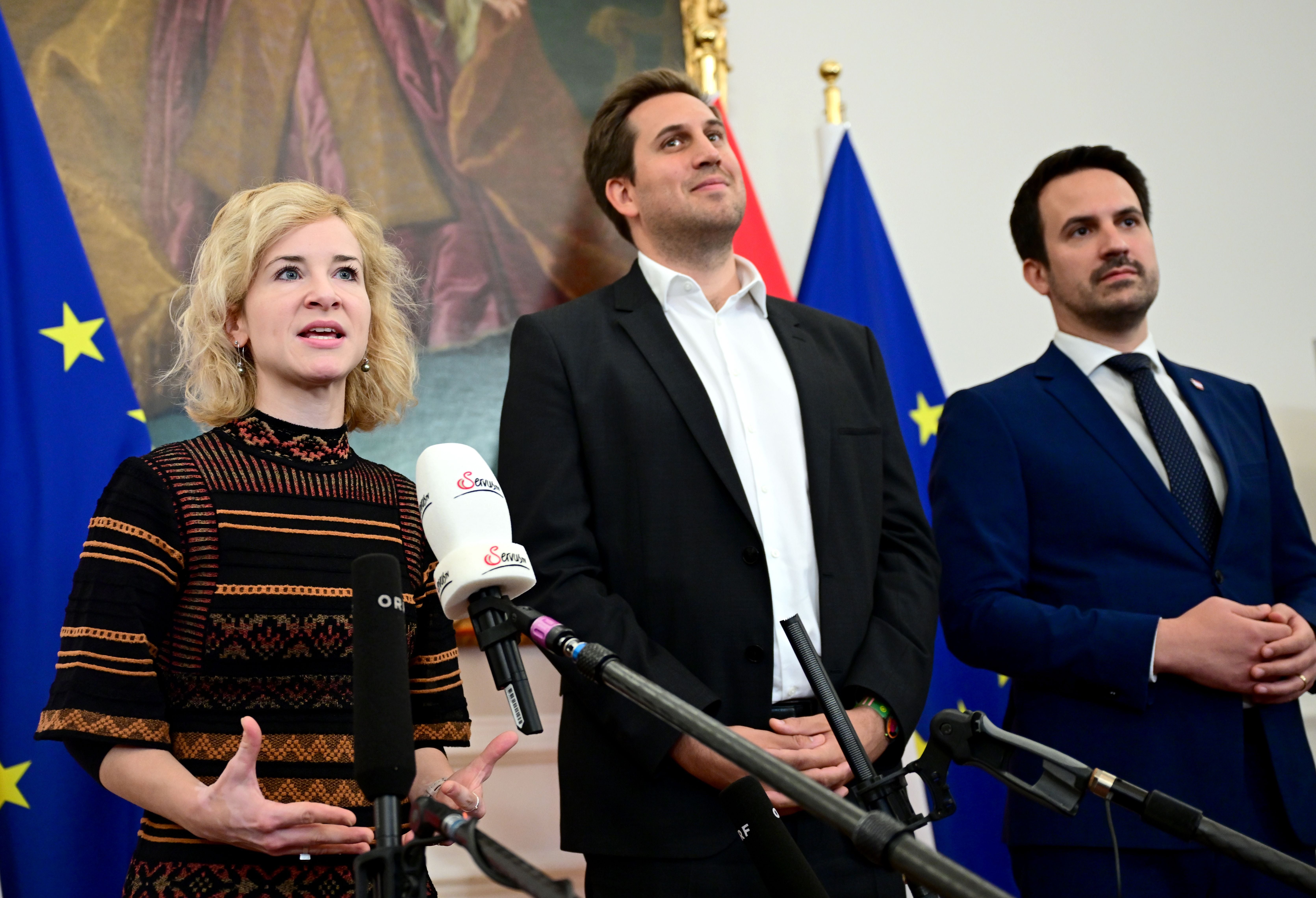 Drei Personen bei einer Pressekonferenz mit Mikrofonen und EU-Flaggen im Hintergrund.