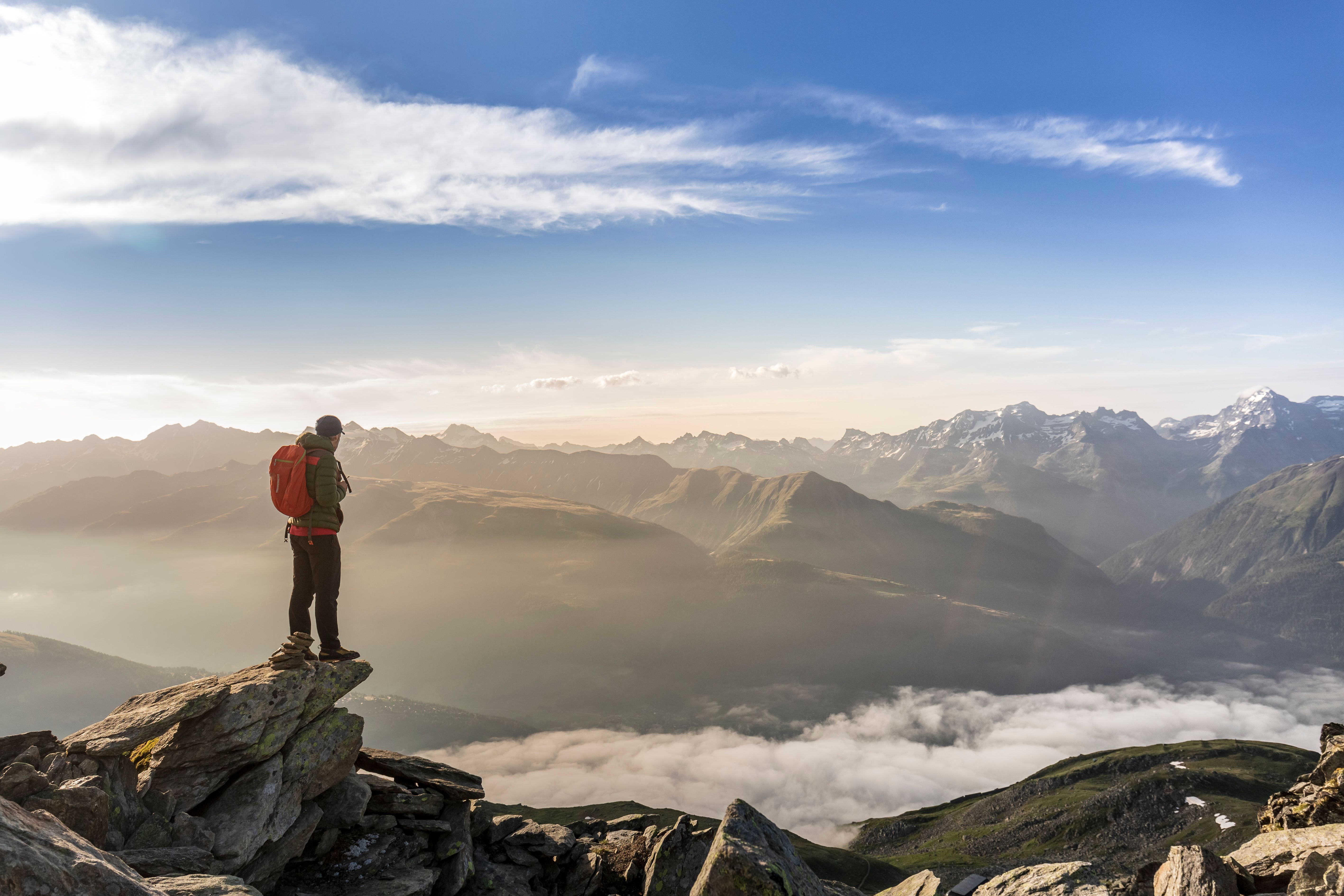Wanderer mit rotem Rucksack steht auf Felsgipfel und blickt über ein weites Bergpanorama.