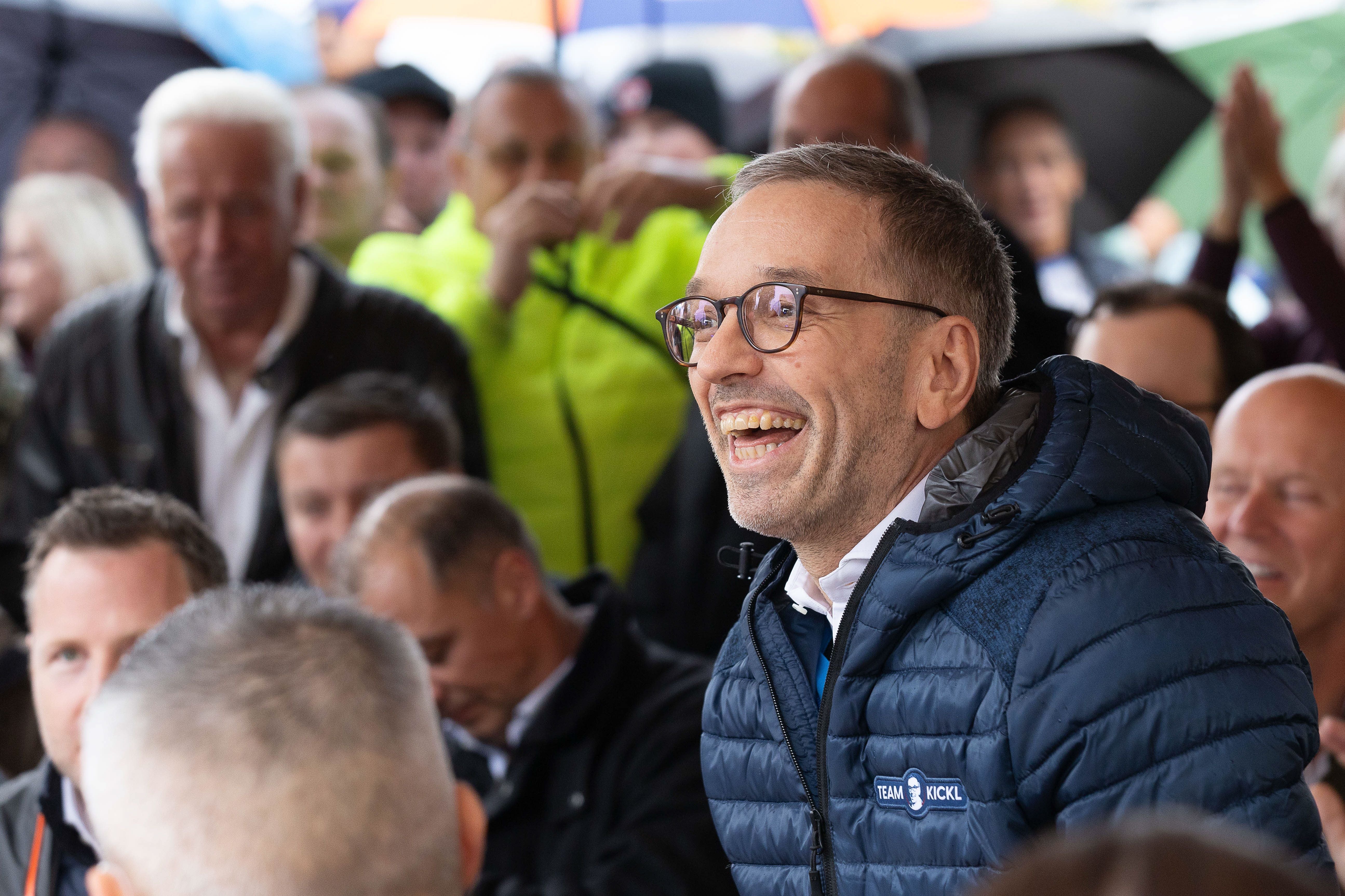 Ein Mann im blauen Steppjacke lacht inmitten einer Menschenmenge bei einer Veranstaltung.