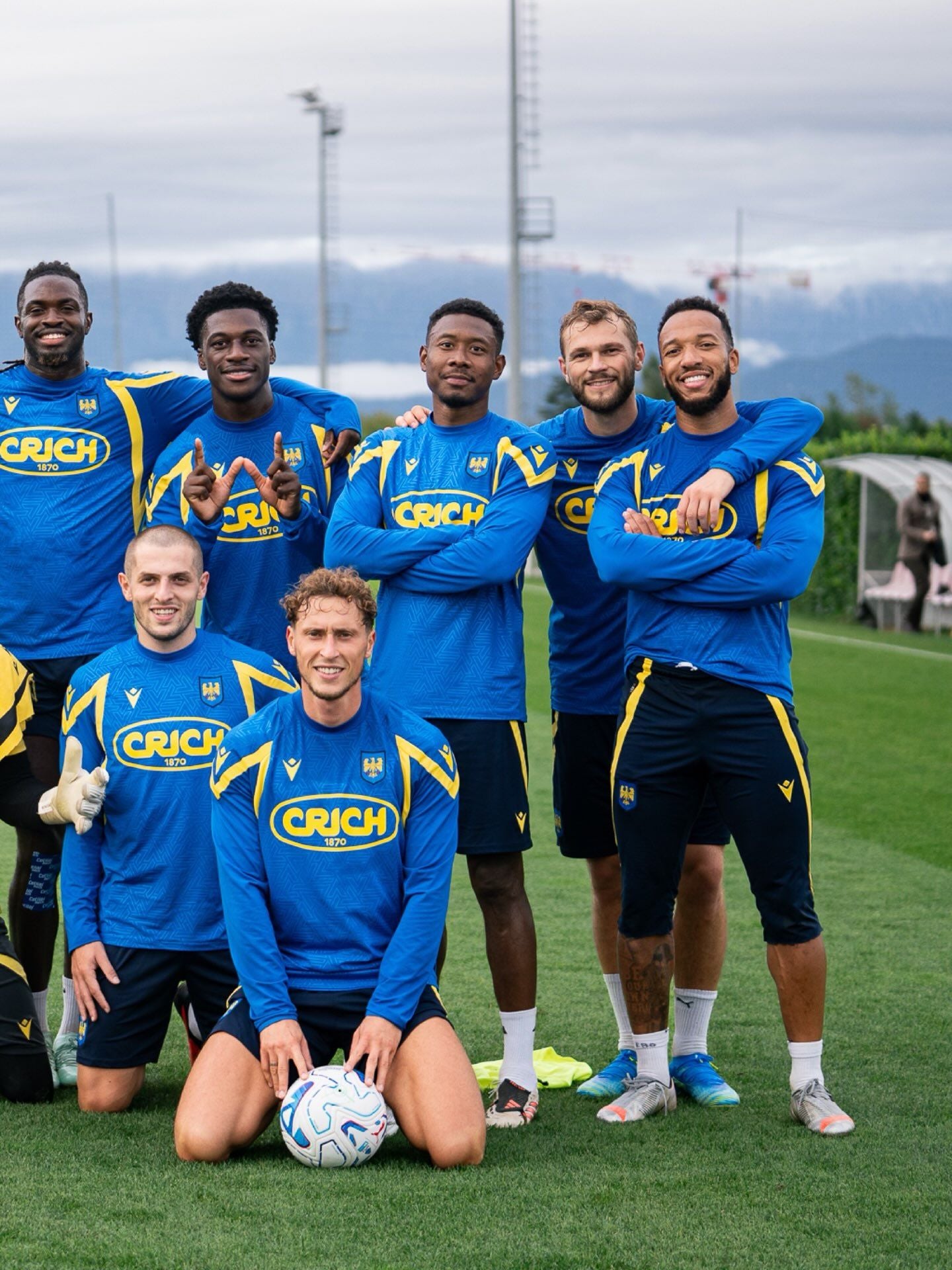 Sieben Fußballspieler posieren lachend im blauen Udinese-Trikot auf einem Trainingsplatz.