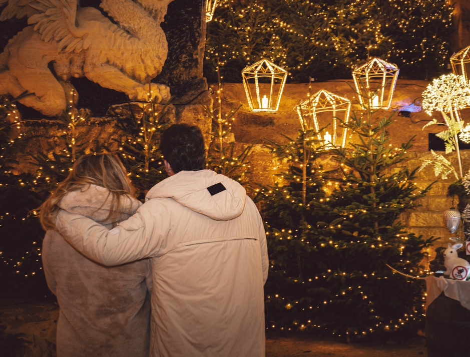 Paar in Wintermänteln steht vor weihnachtlich beleuchteten Tannenbäumen in Valkenburg.