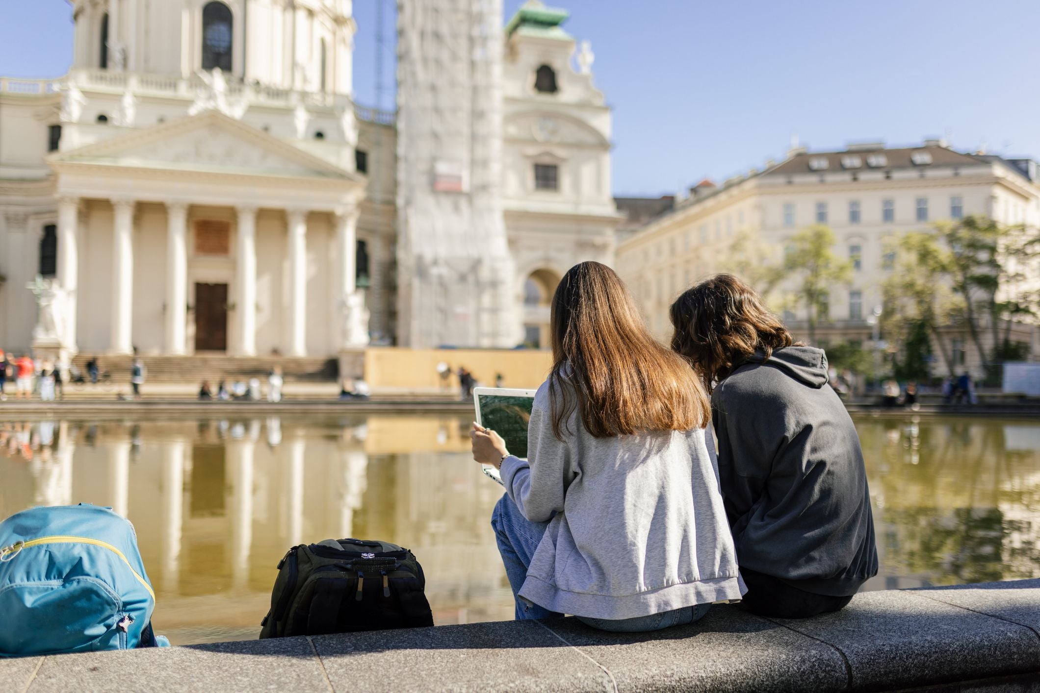 Zwei Jugendliche sitzen vor der Karlskirche in Wien und schauen gemeinsam auf einen Laptop.