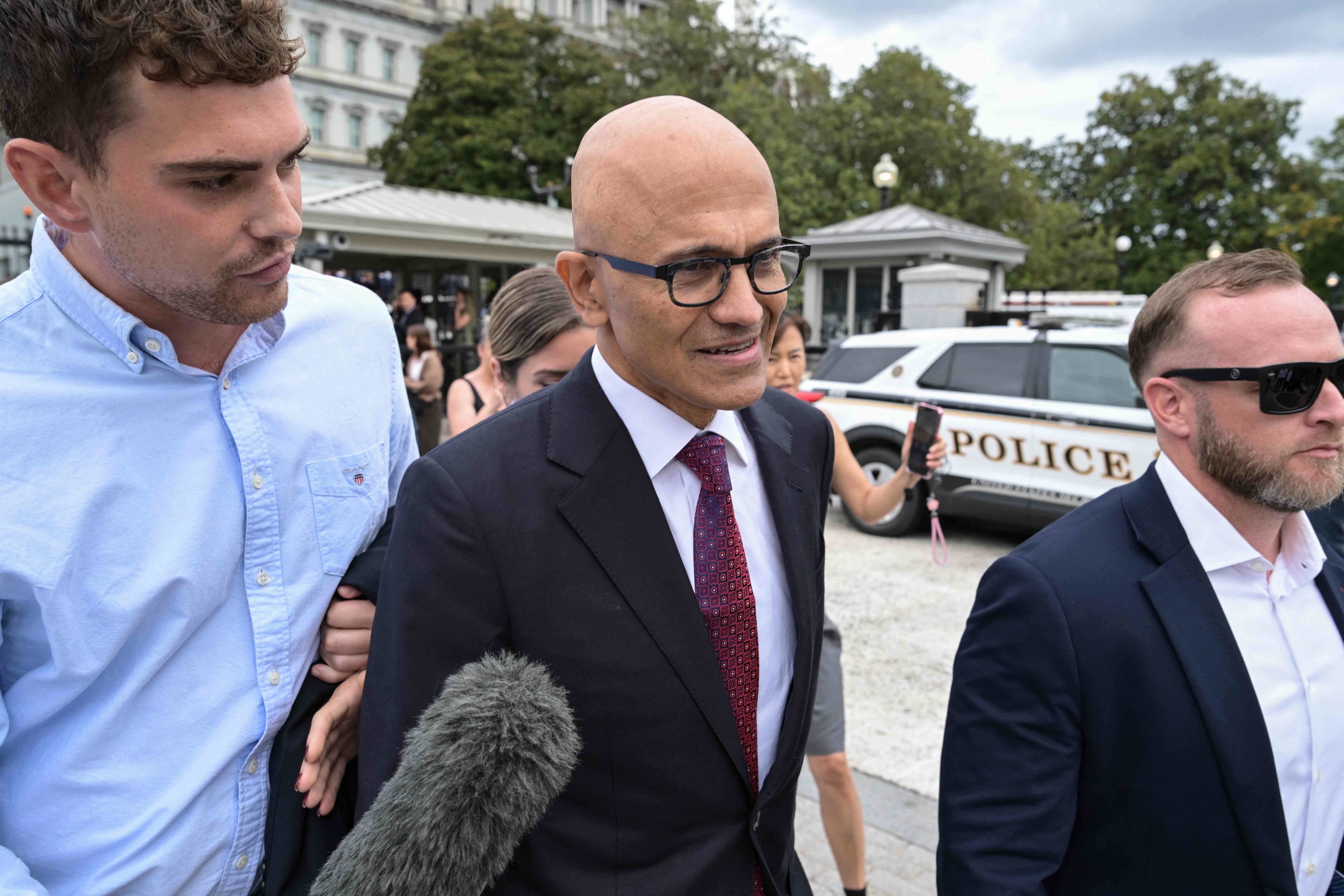 Ein Mann im Anzug verlässt mit Begleitern ein Gebäude, im Hintergrund ein Polizeifahrzeug.
