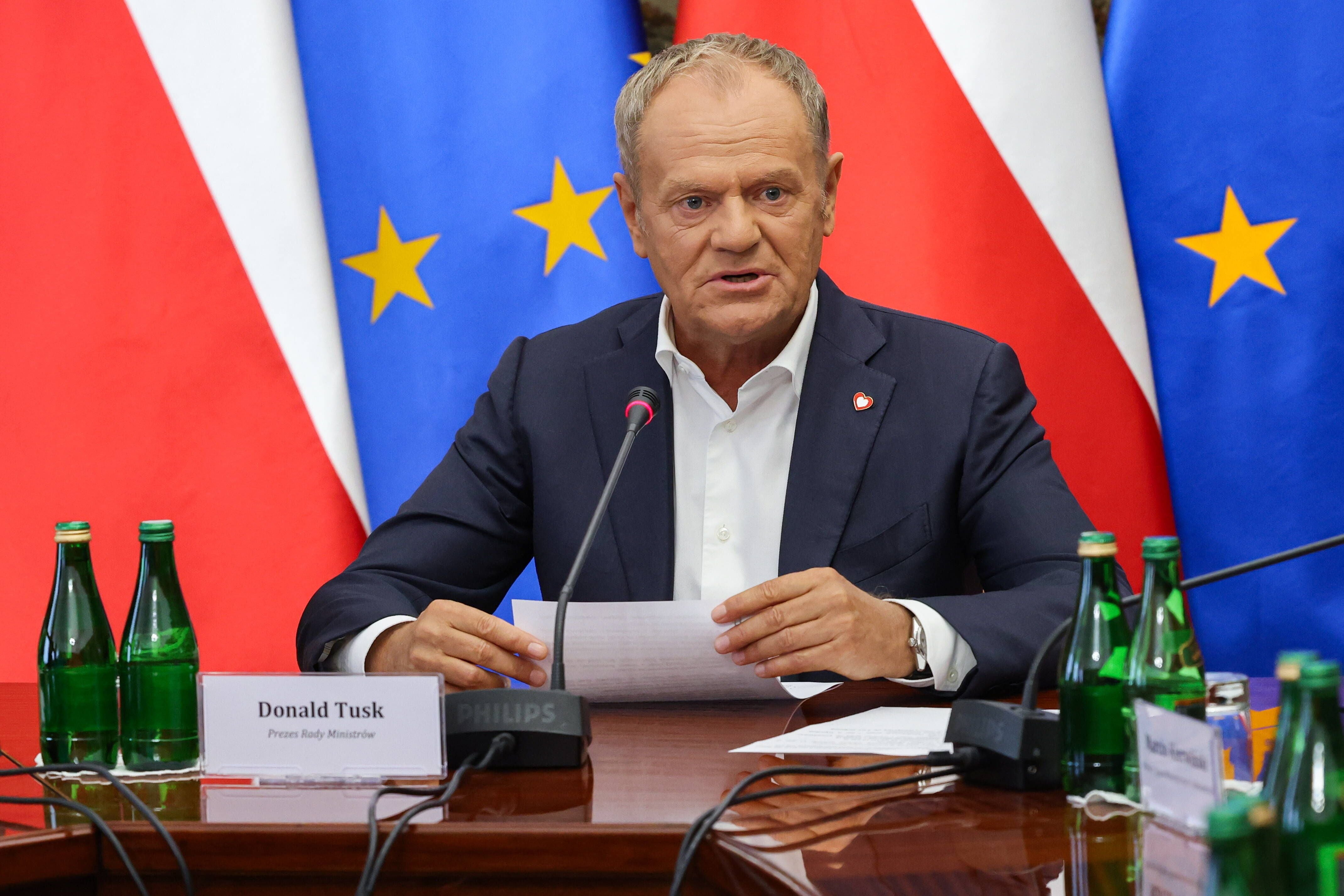 Ein Mann im Anzug sitzt mit Papieren vor Mikrofonen, hinter ihm polnische und EU-Flaggen.