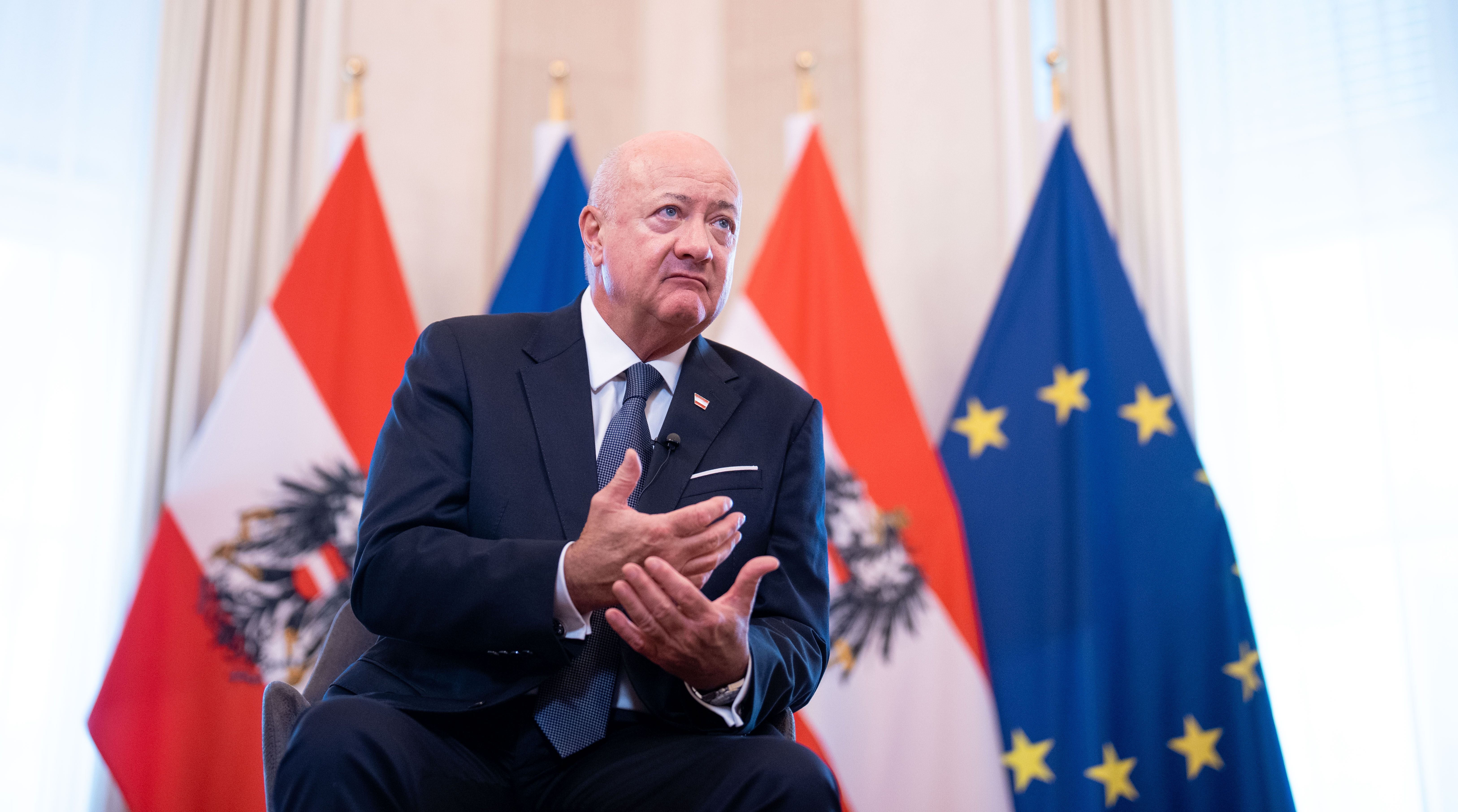 Ein Mann im Anzug spricht vor österreichischen und EU-Flaggen in einem Interviewraum.