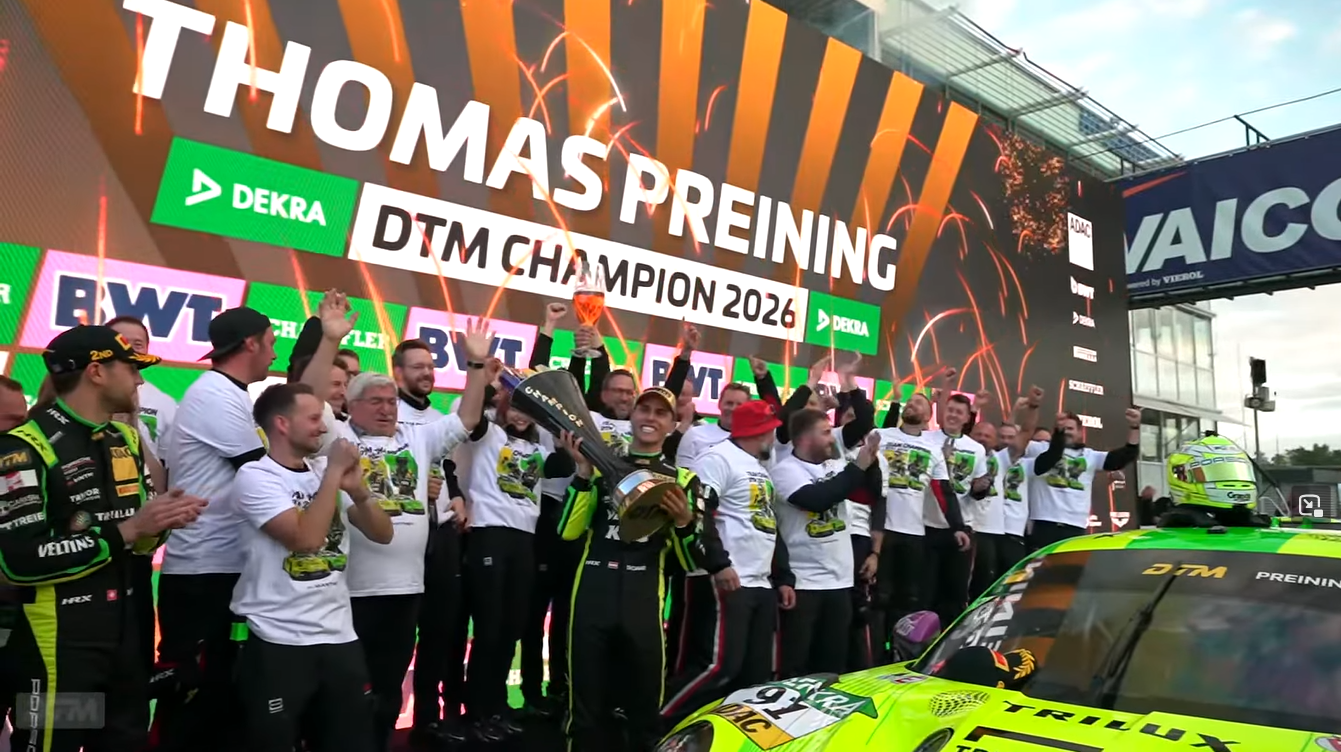 Thomas Preining hält einen Pokal vor seinem jubelnden Team. Neben ihm steht ein neongrüner Rennwagen, dahinter zeigt eine Leinwand "DTM Champion 2026".