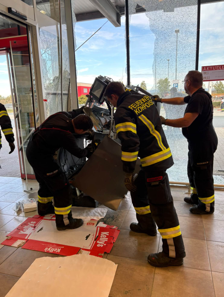 Feuerwehrleute sichern einen gesprengten Bankomaten vor einer zerstörten Glasscheibe.