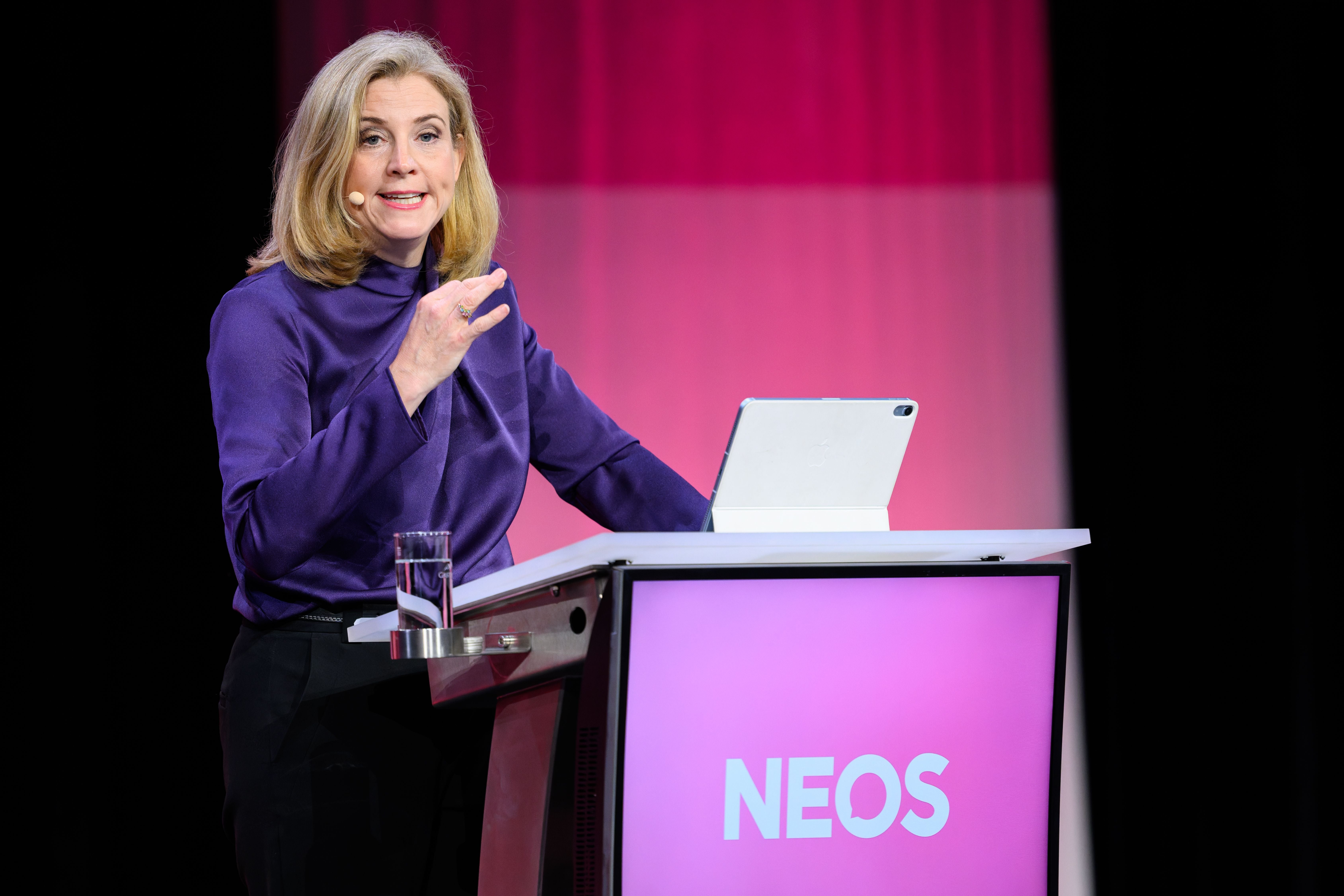 Eine Frau spricht an einem Rednerpult mit NEOS-Logo und Tablet bei einer Veranstaltung.