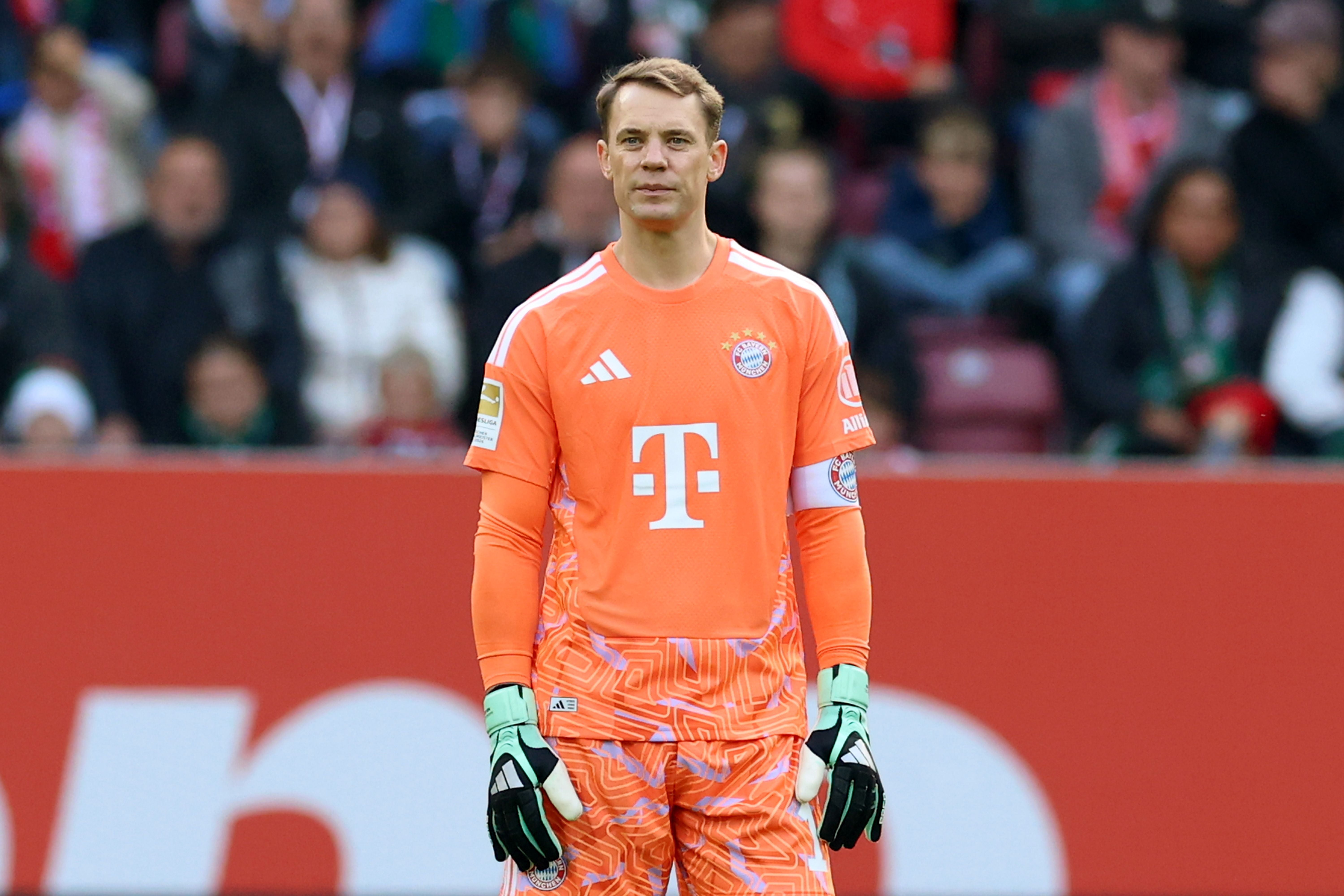 Ein Fußballtorwart im orangefarbenen Trikot steht auf dem Spielfeld, Zuschauer im Hintergrund.