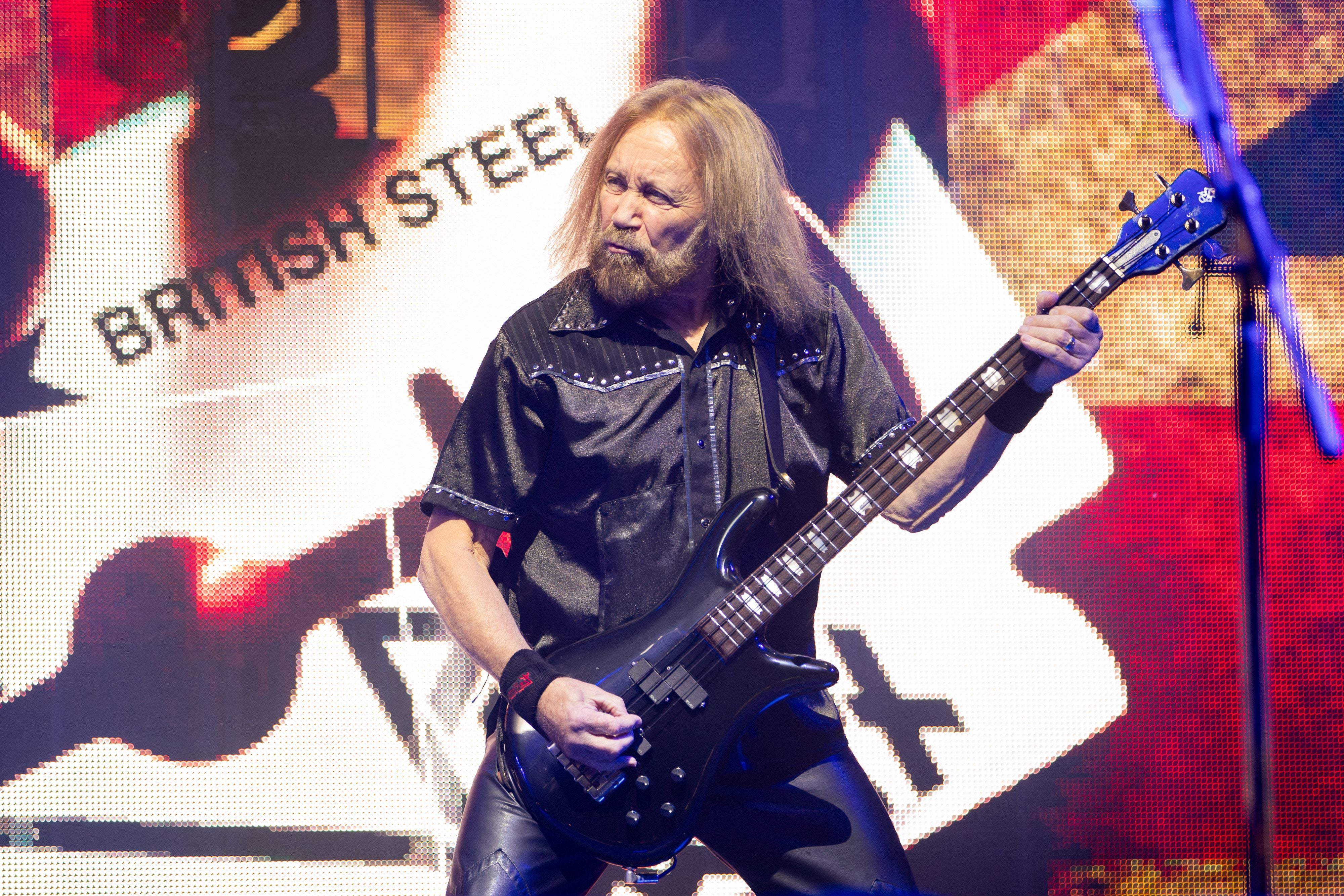 Ein Musiker spielt E-Bass auf einer Bühne mit "British Steel"-Hintergrundgrafik.