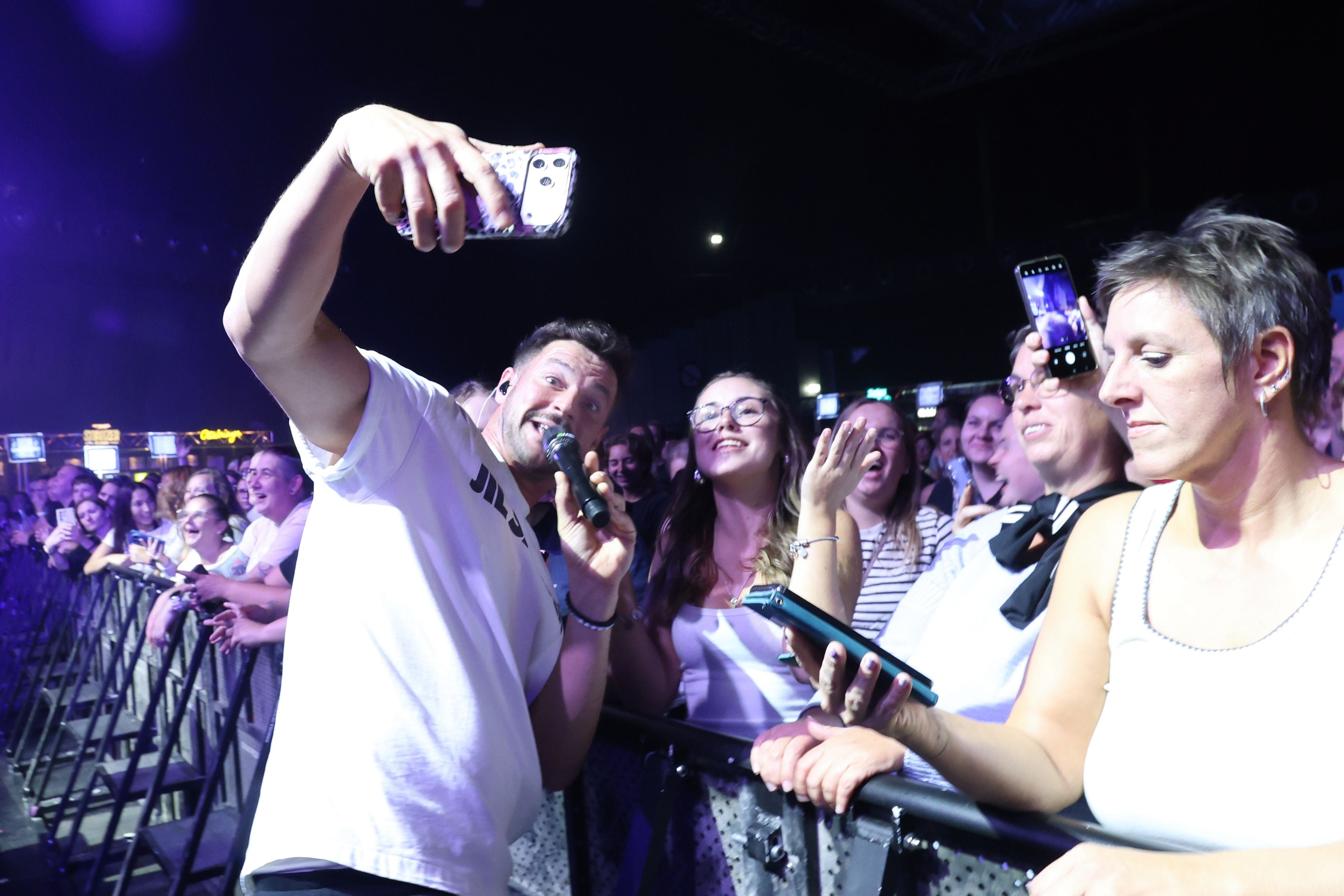 Ein Sänger macht ein Selfie mit begeisterten Fans bei einem Konzert in einer Halle.