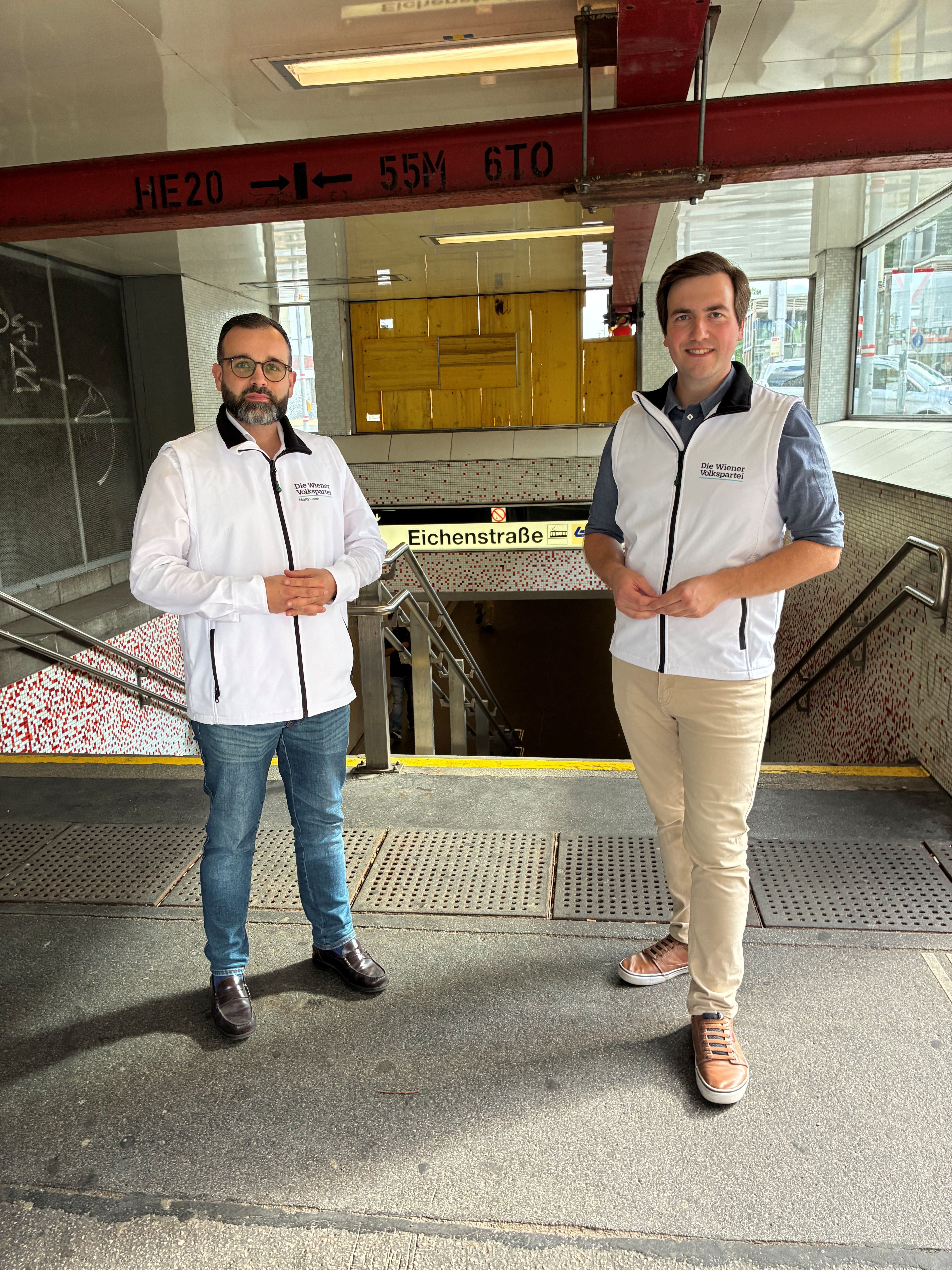 Zwei Männer in weißen ÖVP-Westen stehen vor einem U-Bahn-Eingang "Eichenstraße".