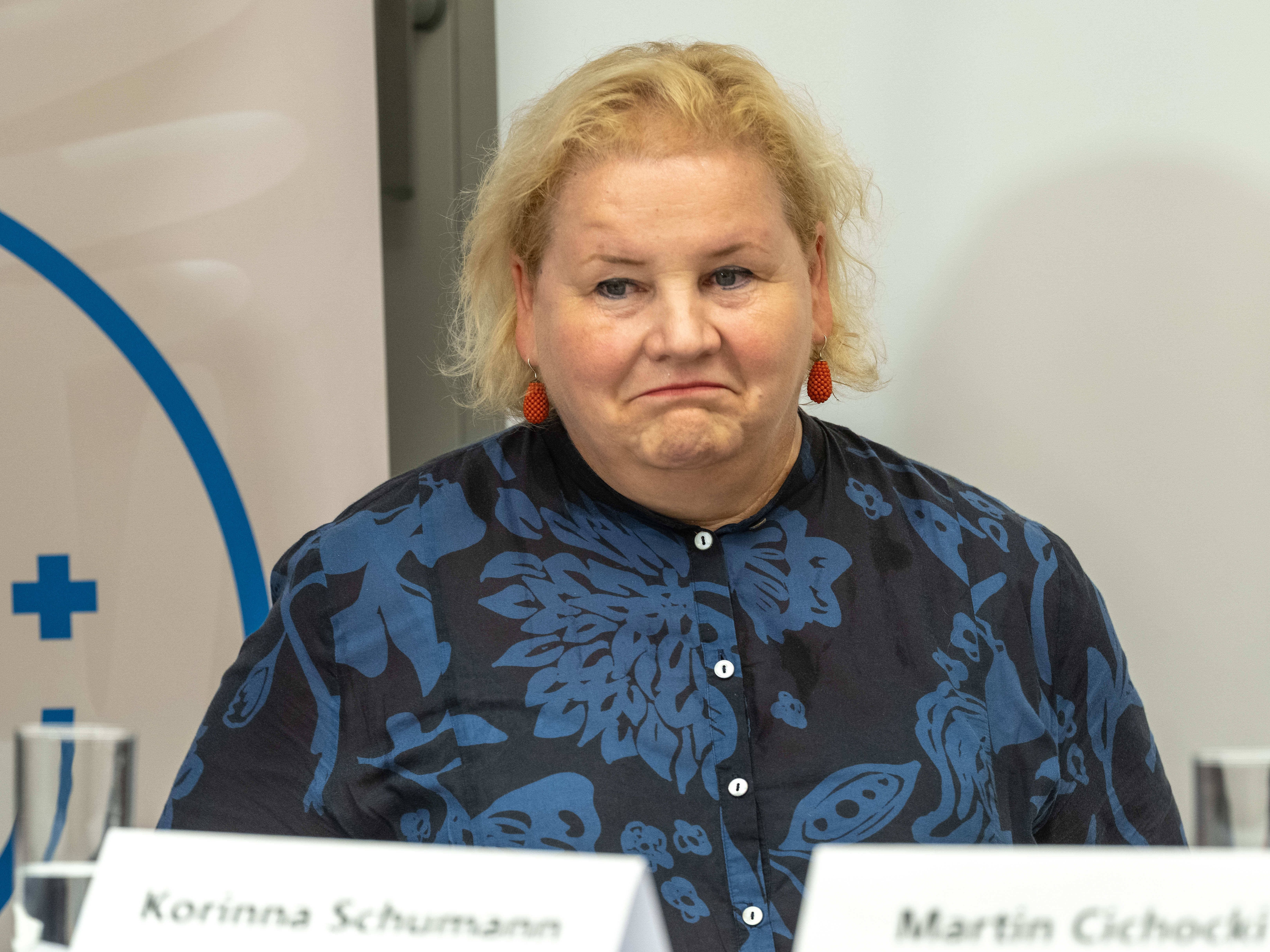 Frau mit blondem Haar und gemustertem Hemd bei einer Pressekonferenz in einem Konferenzraum.