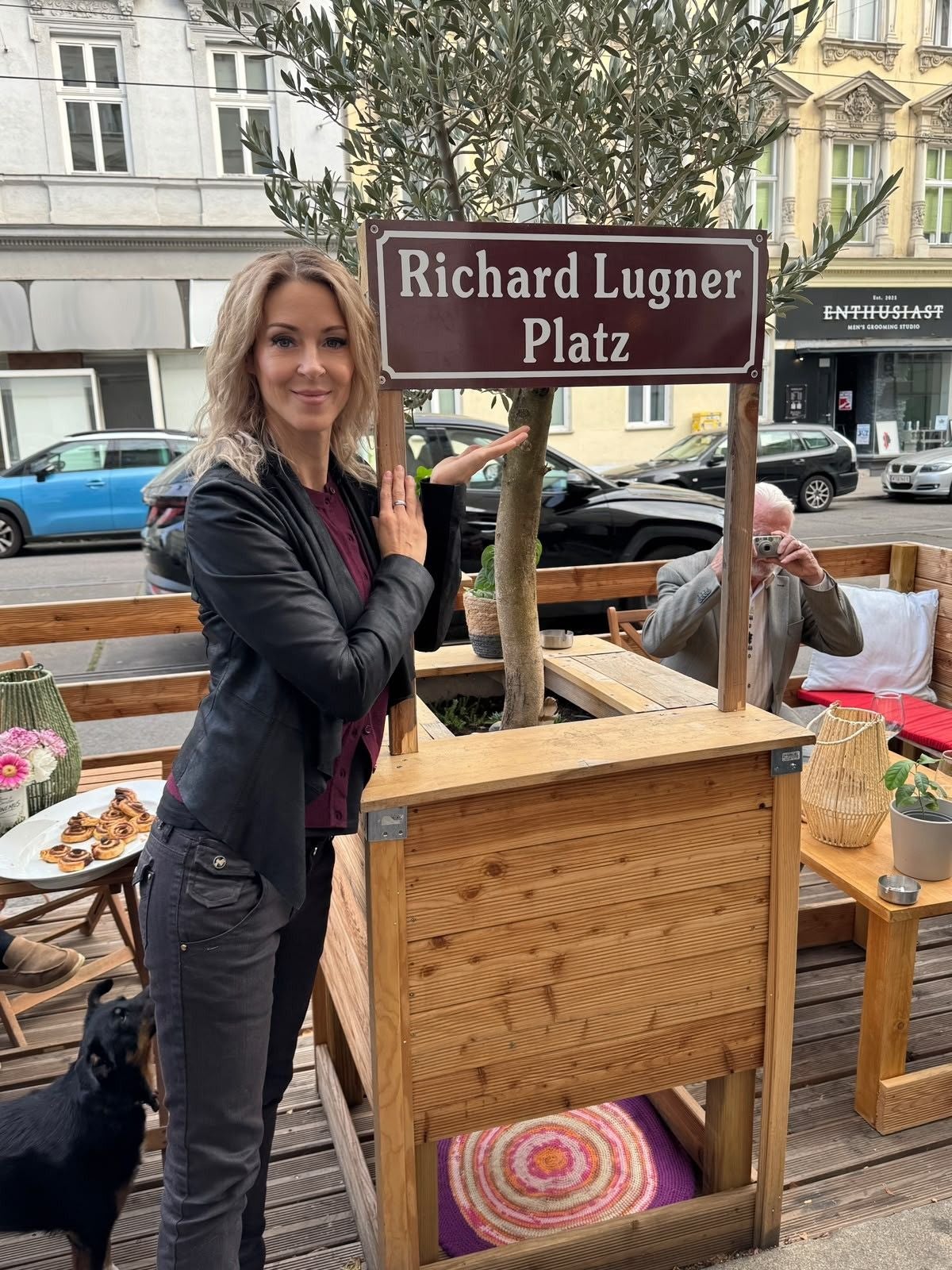 Frau steht neben einem Schild mit der Aufschrift "Richard Lugner Platz" auf einer Terrasse.