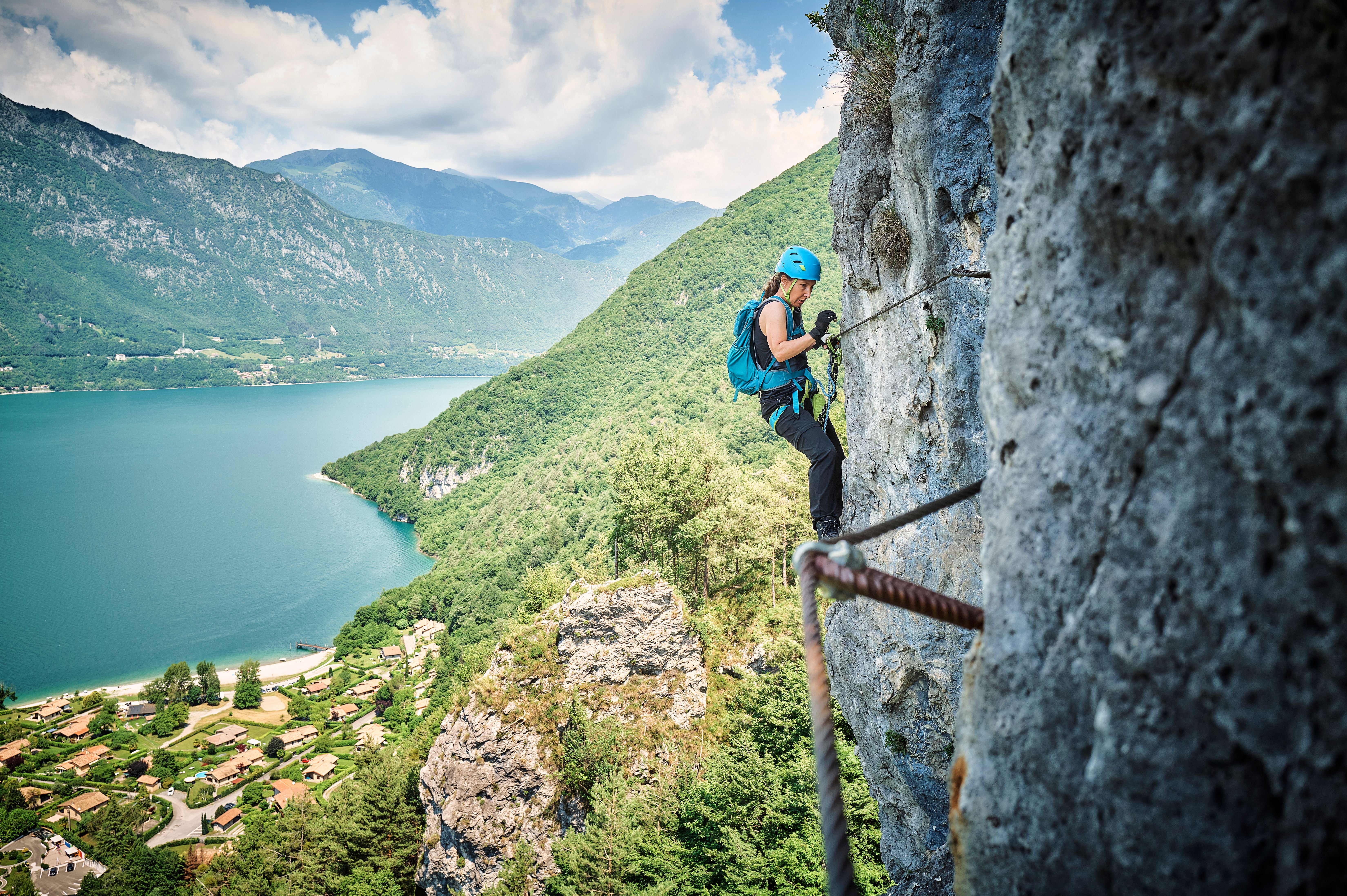 Eine Frau klettert gesichert an einer Felswand mit Blick auf einen See und grüne Berge.