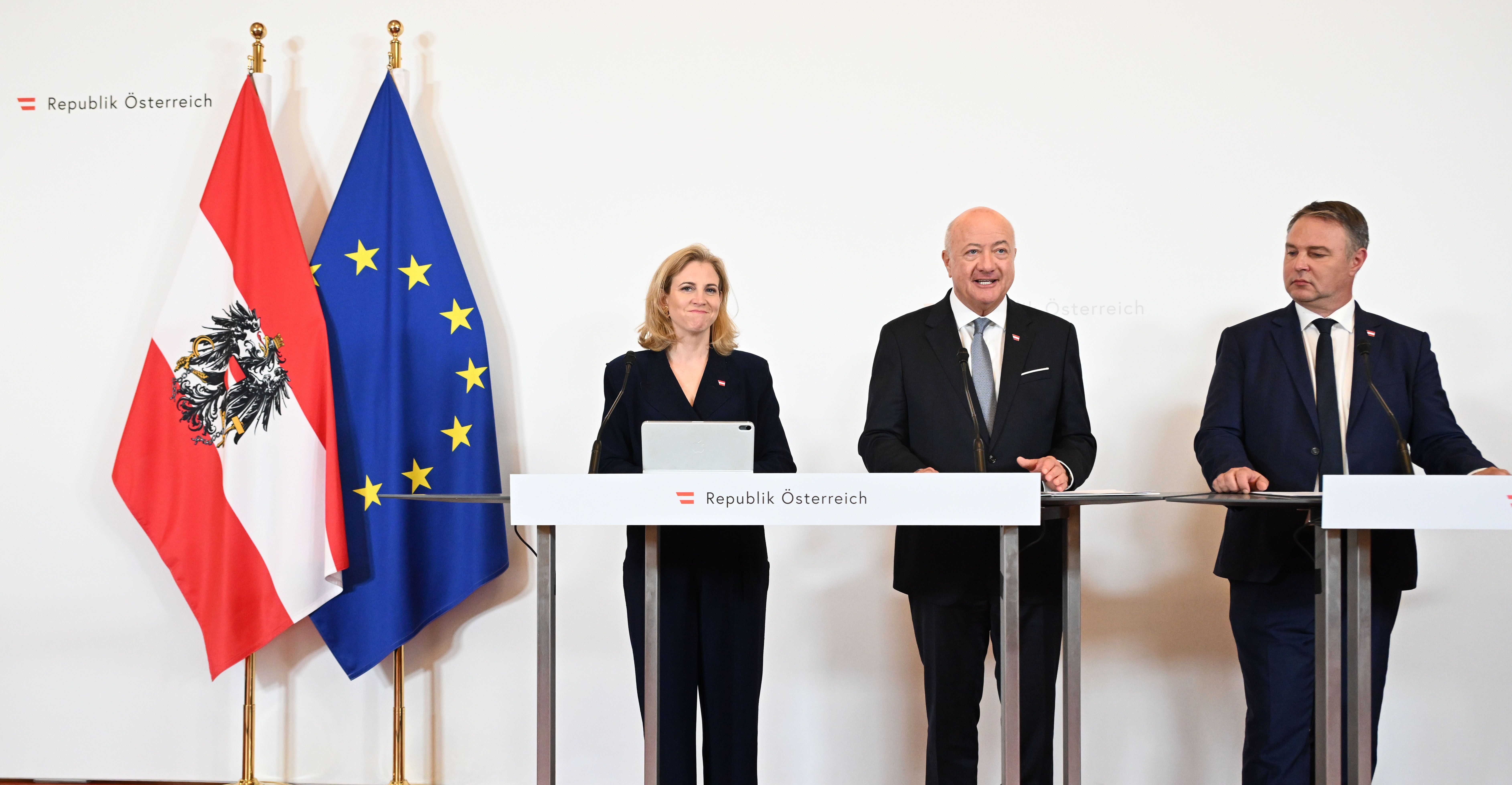 Drei Politiker bei einer Pressekonferenz mit österreichischer und EU-Flagge im Hintergrund.