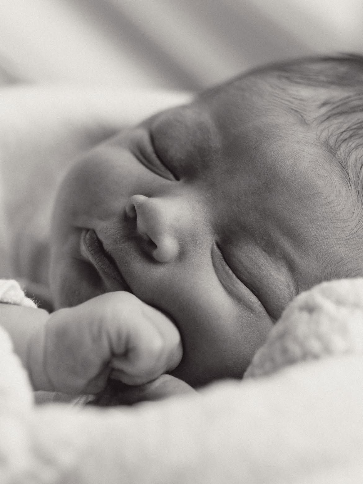 Schwarzweißaufnahme eines schlafenden Babys mit geschlossener Faust am Gesicht.