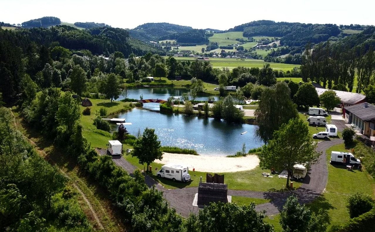 Campingplatz mit Wohnmobilen und See in grüner, hügeliger Landschaft bei sonnigem Wetter.