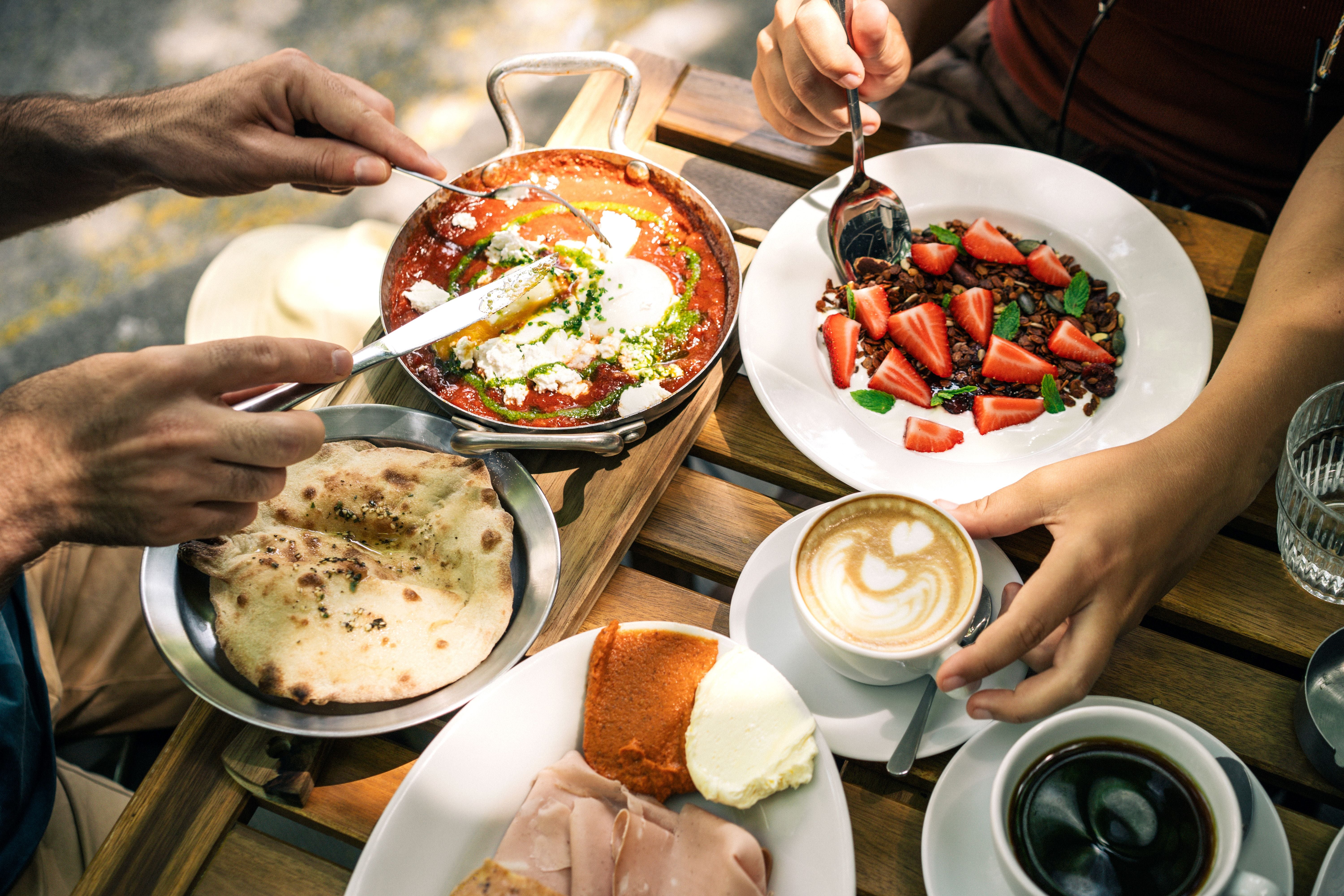 Brunch mit Shakshuka, Fladenbrot, Müsli mit Erdbeeren und Cappuccino an einem Holztisch im Freien.